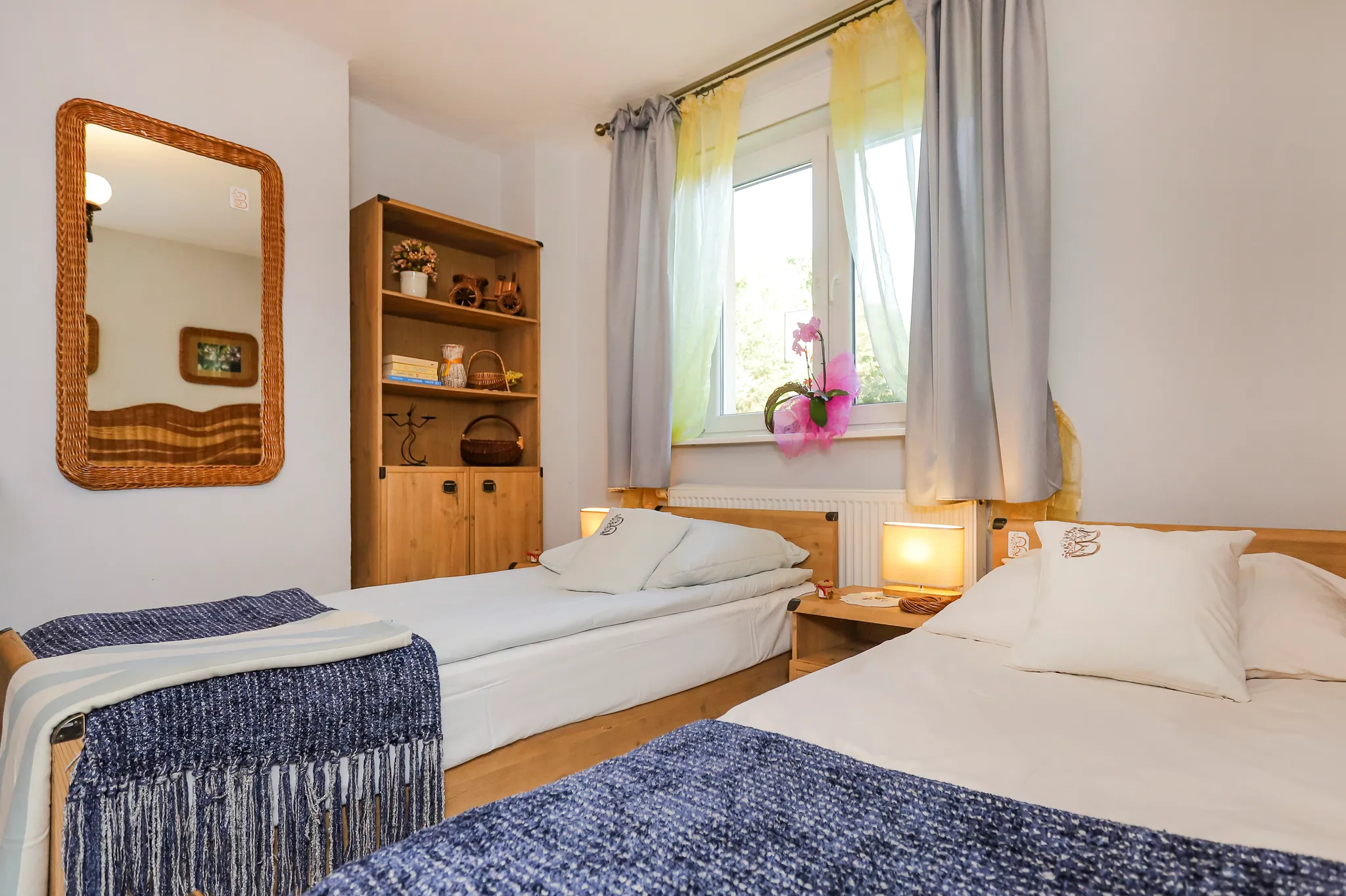 Przytulna sypialnia z łóżkiem, żółtymi zasłonami, telewizorem na komodzie oraz wiklinowymi meblami przy ścianie z czerwonej cegły.