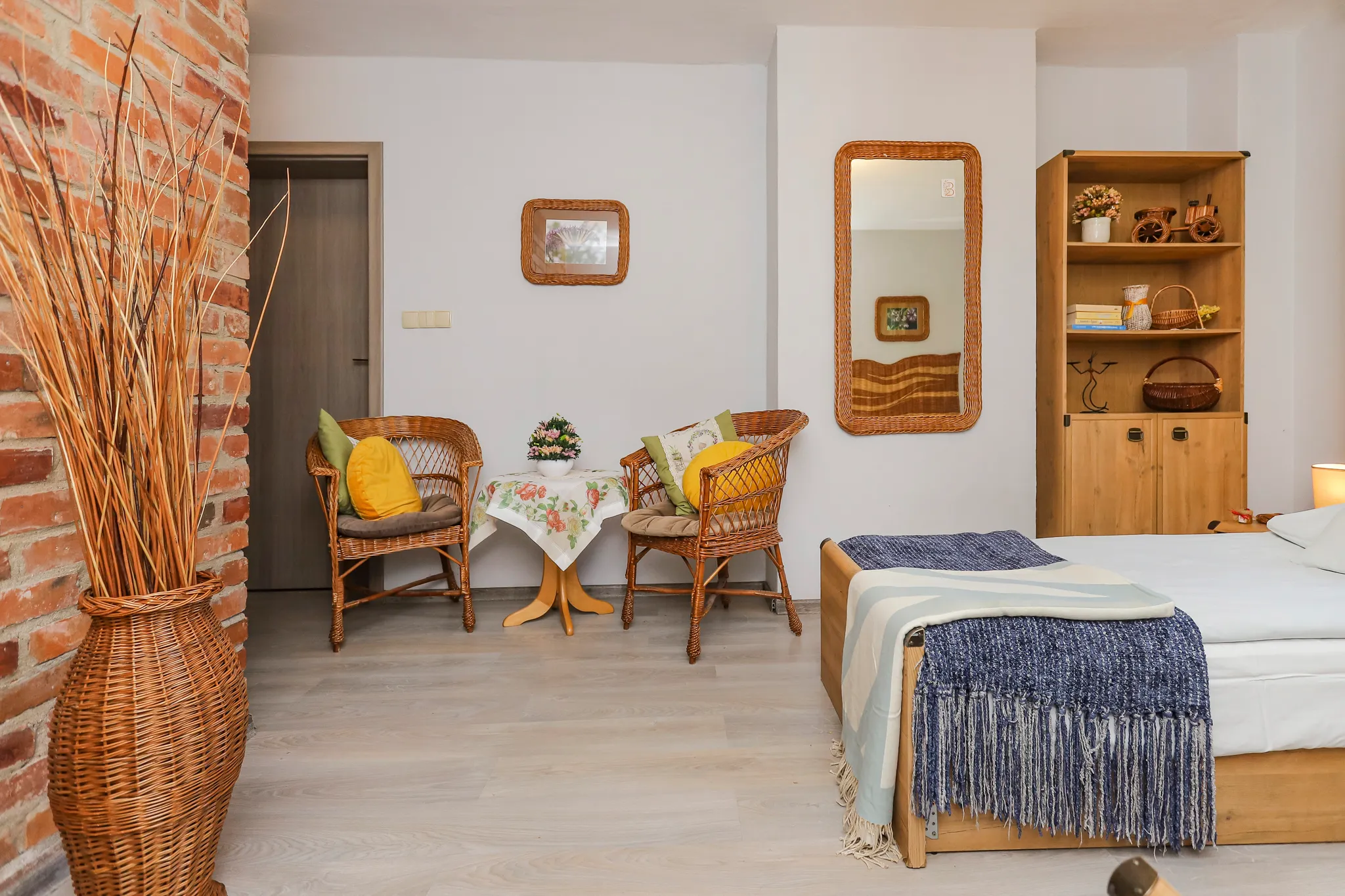 Przytulna sypialnia z łóżkiem, żółtymi zasłonami, telewizorem na komodzie oraz wiklinowymi meblami przy ścianie z czerwonej cegły.