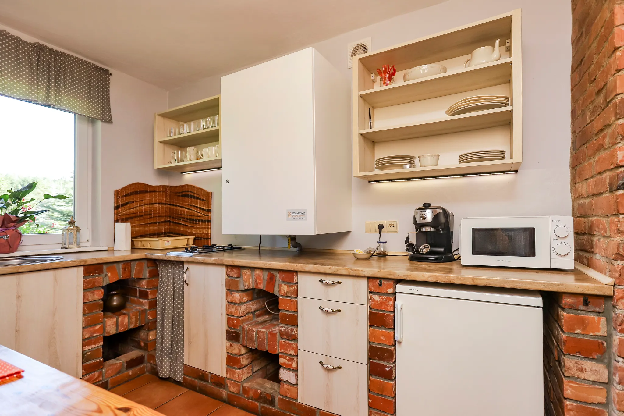 Kuchnia z ceglaną ścianą, drewnianymi blatami, białą zastawą na stole i roślinami na parapecie pod oknem.