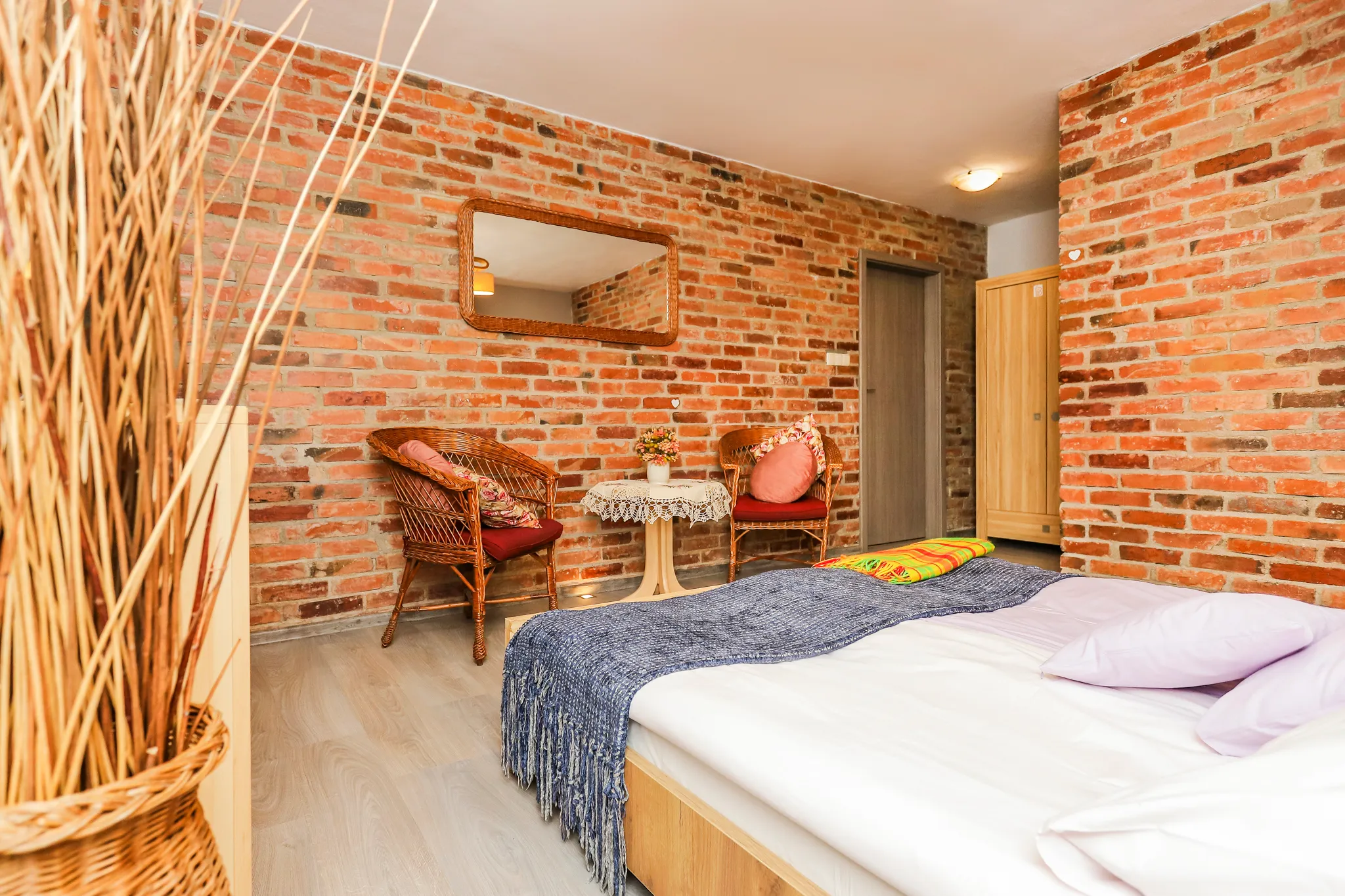 Przytulna sypialnia z łóżkiem, żółtymi zasłonami, telewizorem na komodzie oraz wiklinowymi meblami przy ścianie z czerwonej cegły.