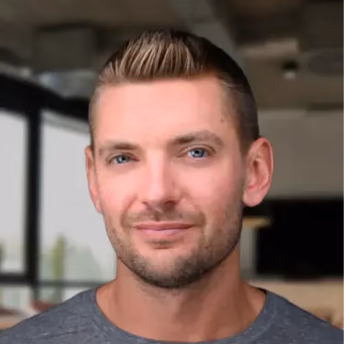 Headshot of a man with short light brown hair and blue eyes, wearing a gray shirt, in an indoor setting.