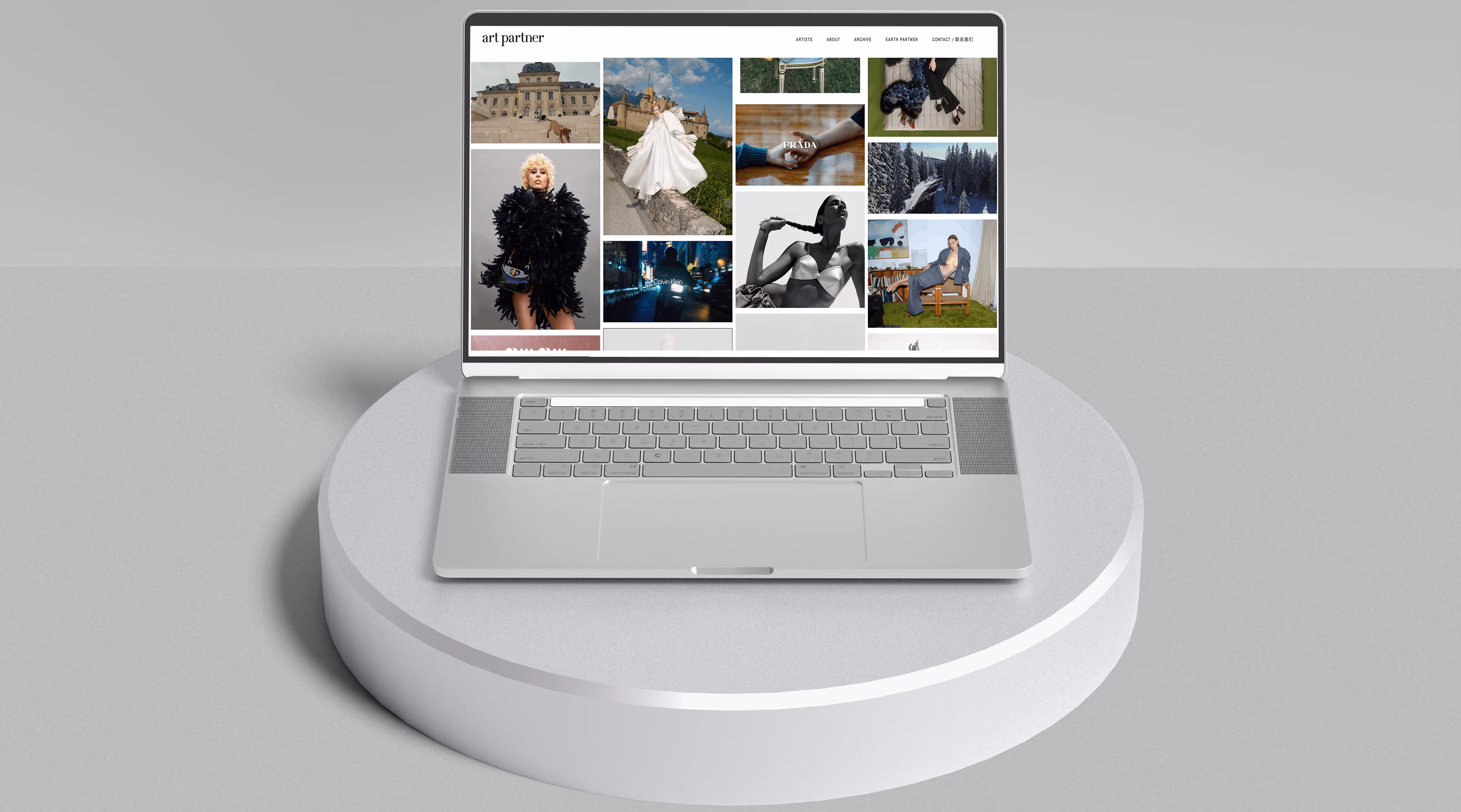 Silver laptop displaying a website named 'art partner' with a grid of fashion and landscape photographs on a white circular pedestal against a gray background.