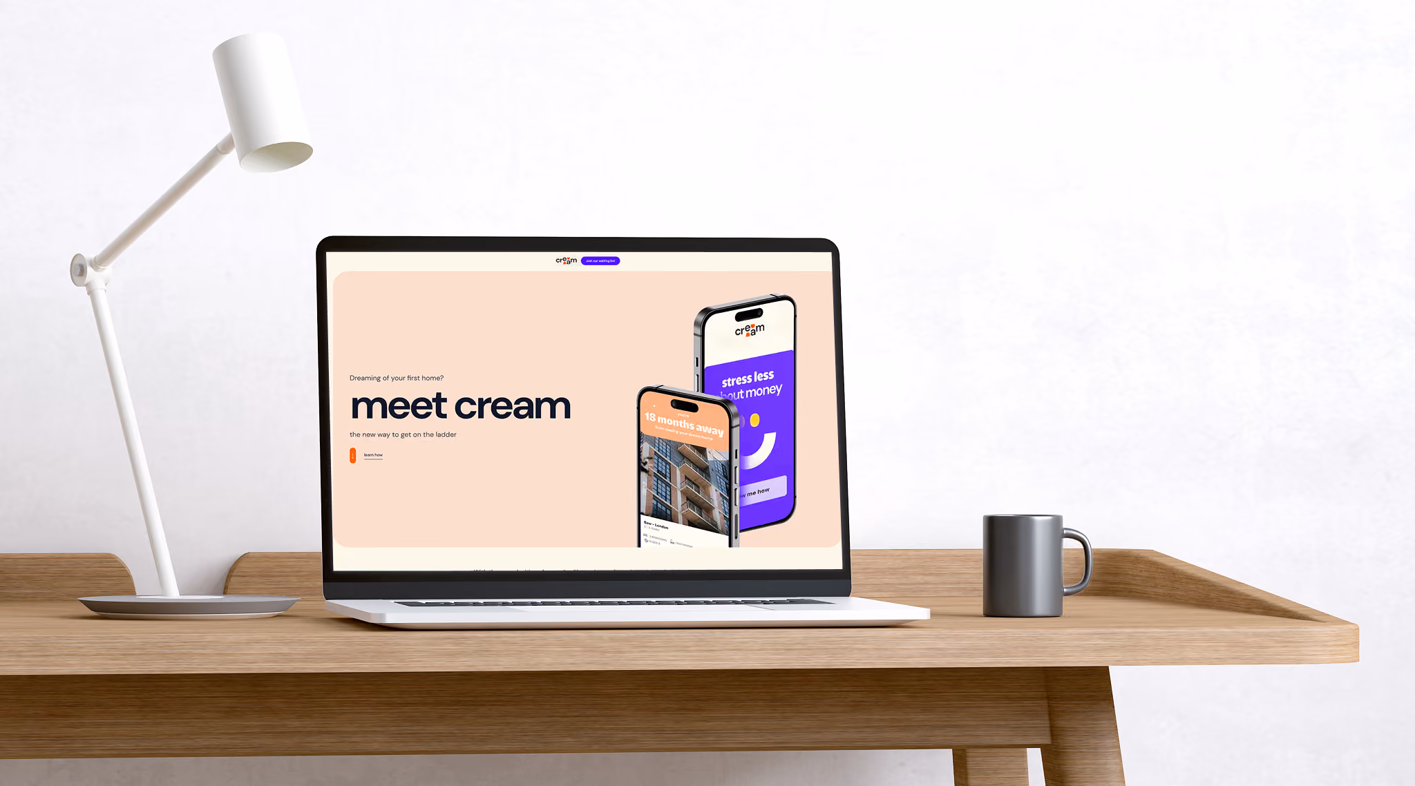 Laptop on wooden desk displaying a website with the heading 'meet cream' and two phone mockups showing home buying app screens, alongside a white desk lamp and gray mug.