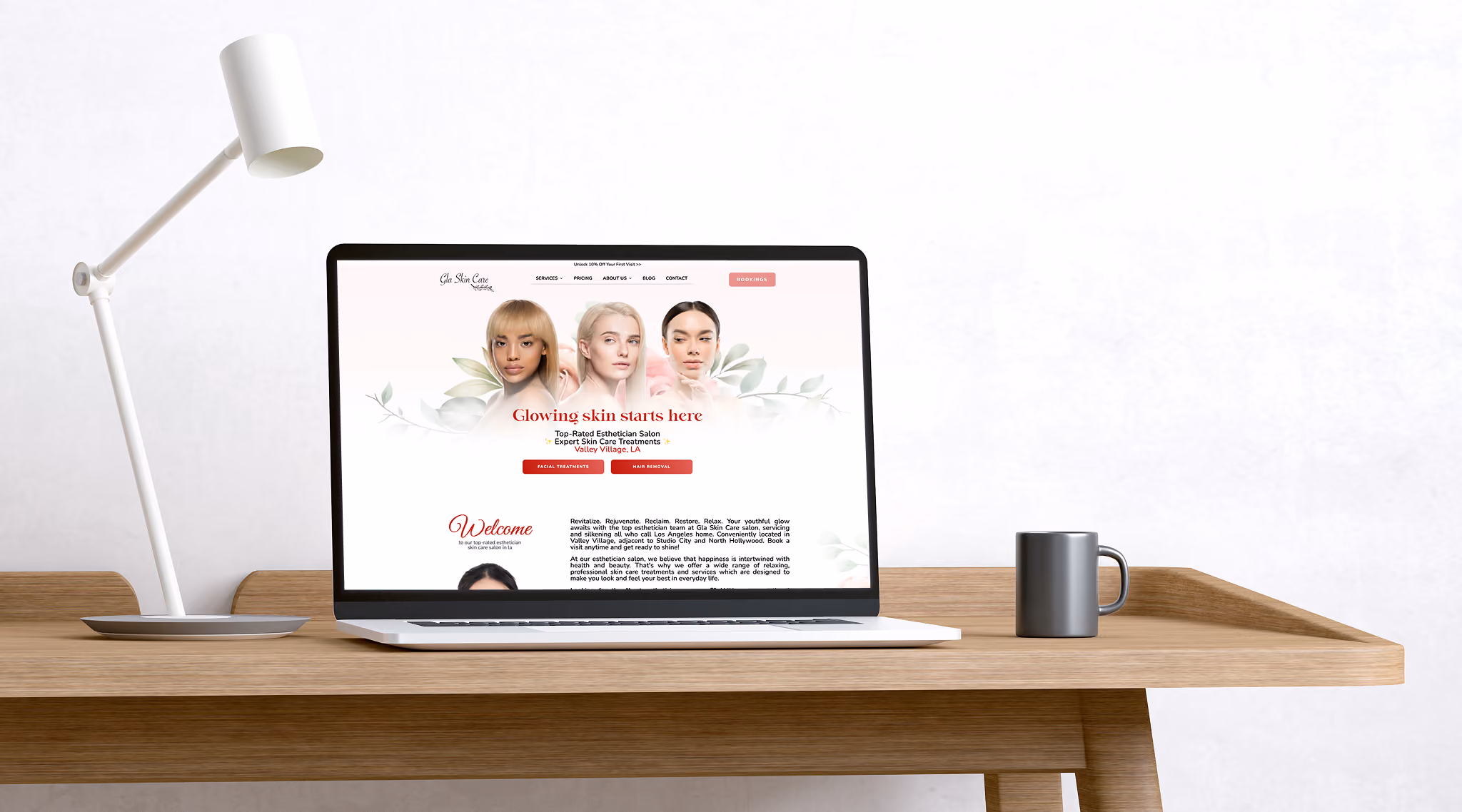Laptop on a wooden desk displaying Gla Skin Care website homepage with images of three women and text about skin treatments, next to a white desk lamp and gray mug.
