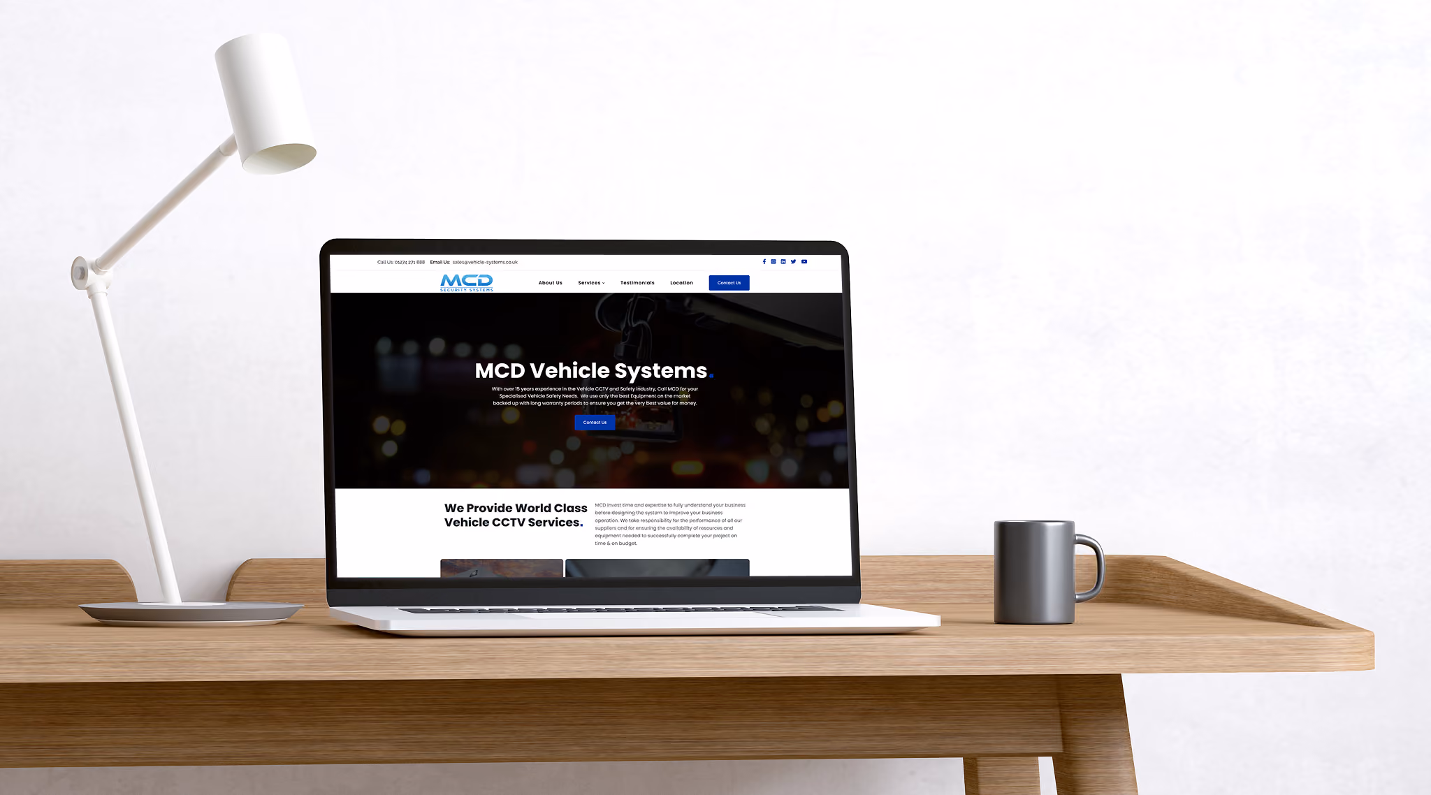 Laptop on wooden desk displaying MCD Vehicle Systems website homepage, with a white desk lamp on the left and a gray mug on the right.