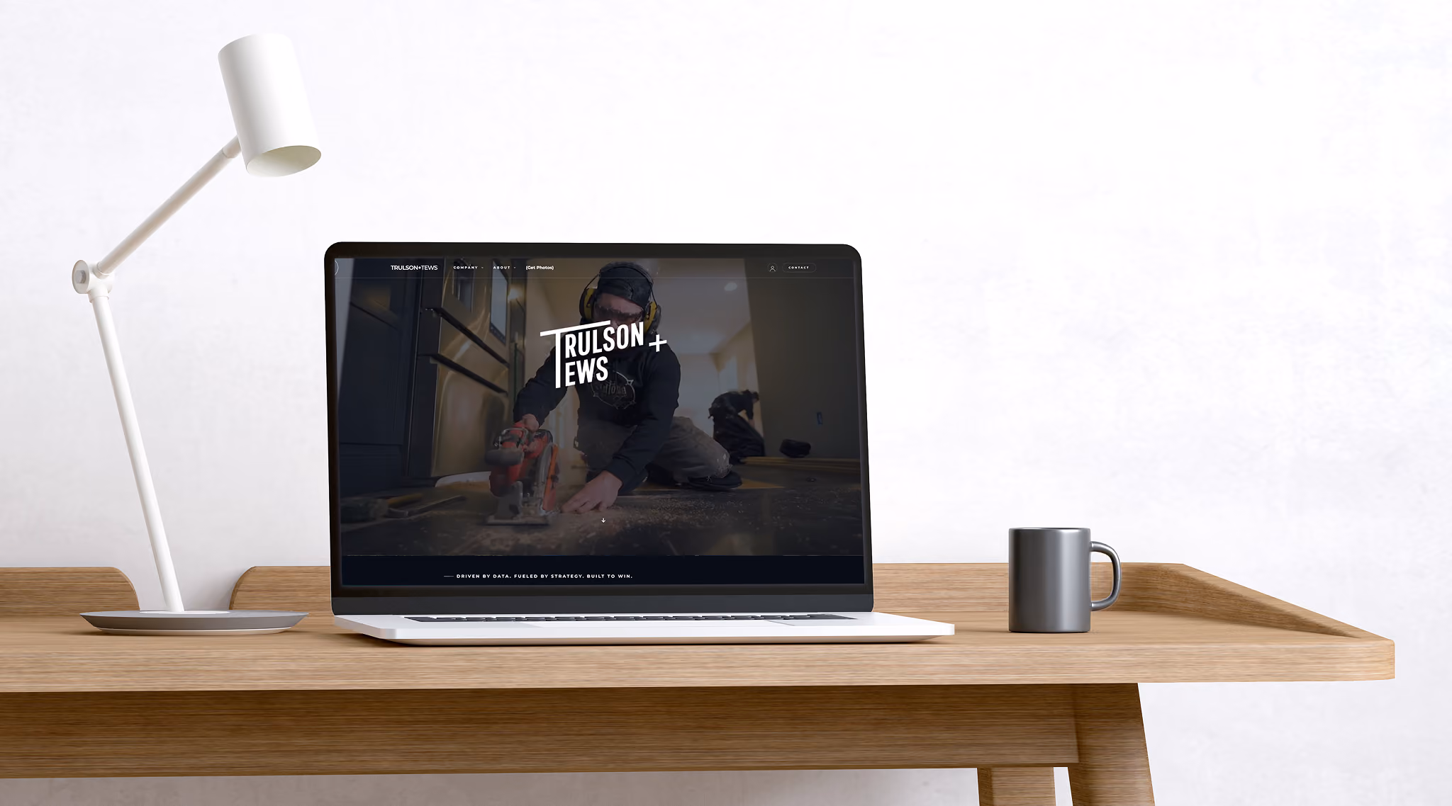 A laptop on a wooden desk displays a website featuring a man using a power saw indoors, flanked by a white desk lamp and a gray coffee mug.