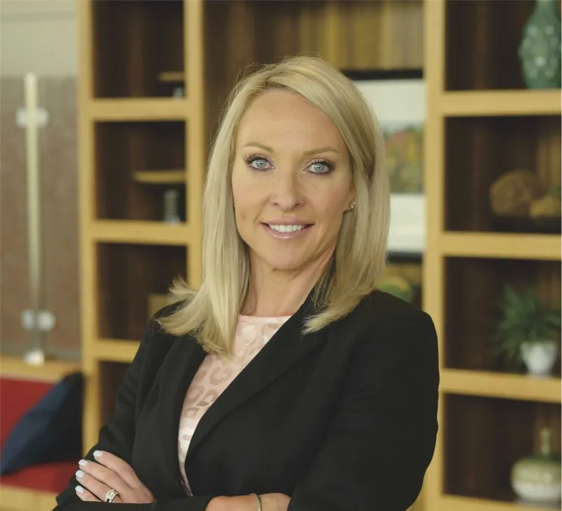Professional woman with blonde hair wearing a black blazer, standing with arms crossed in an office setting with shelves in the background.