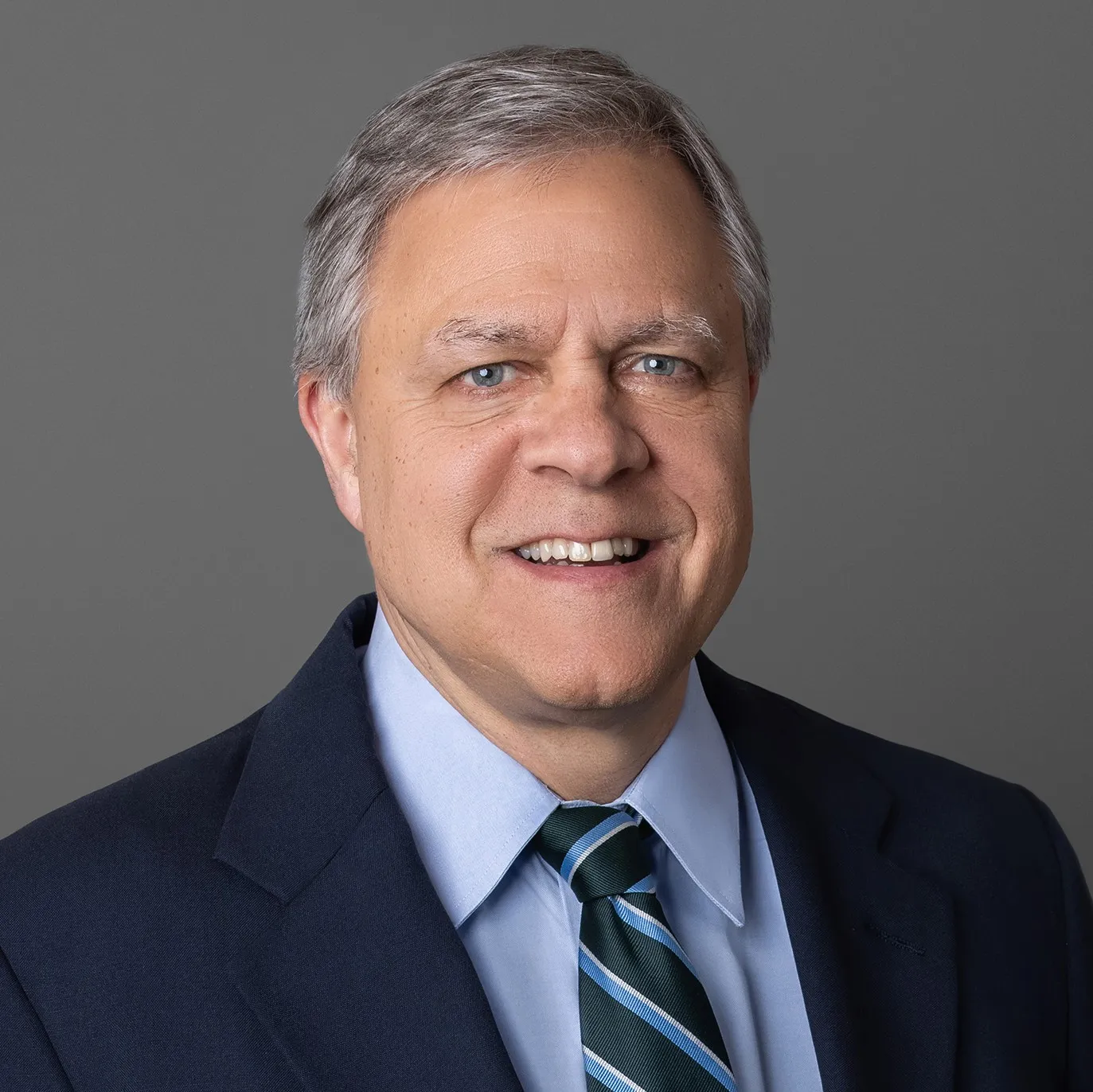 Middle-aged man with gray hair wearing a navy suit, light blue shirt, and striped tie smiling against a gray background.