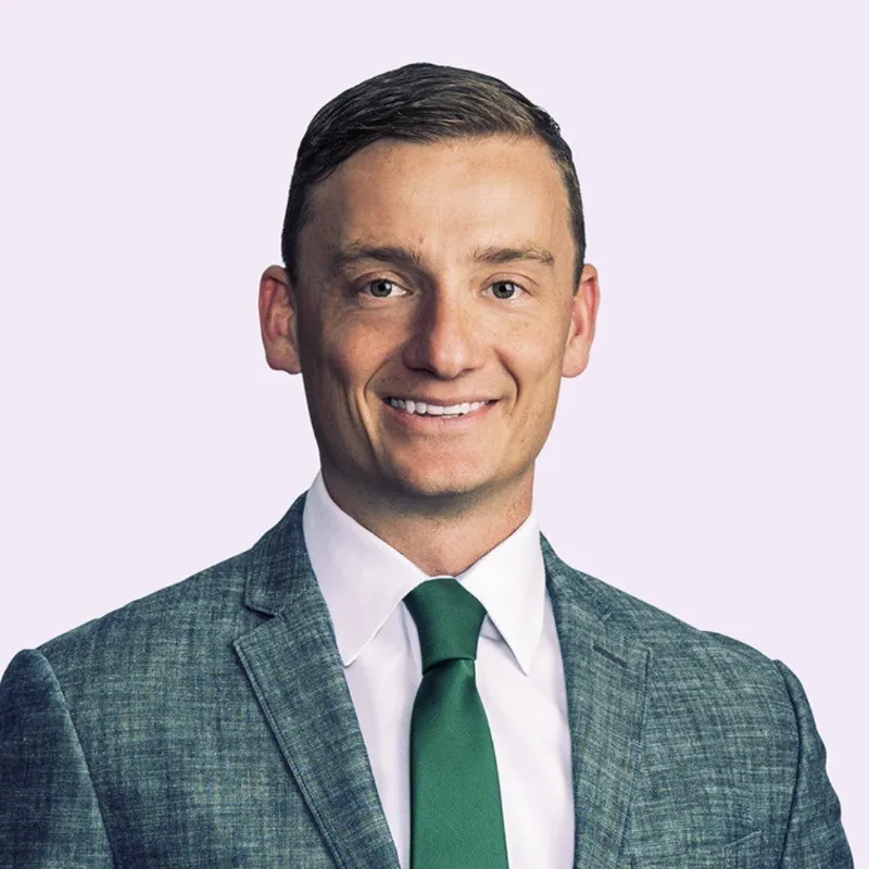 Smiling man in a gray suit jacket, white shirt, and green tie against a light background.