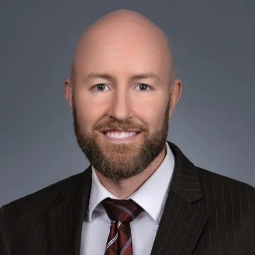 Headshot of a bald man with a beard wearing a dark pinstripe suit, white shirt, and striped tie, smiling against a gray background.