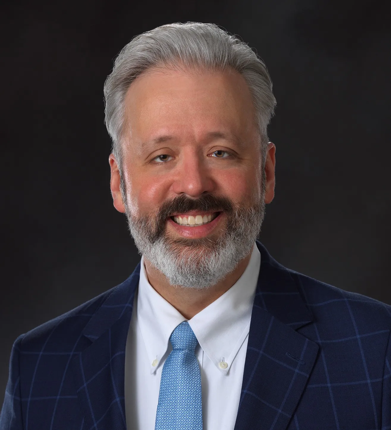 Middle-aged man with gray hair and beard wearing a blue checkered suit, white shirt, and light blue tie smiling against a dark background.