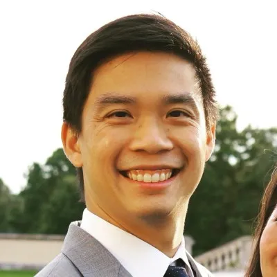 Smiling man with short black hair wearing a gray suit and tie outdoors with trees in the background.