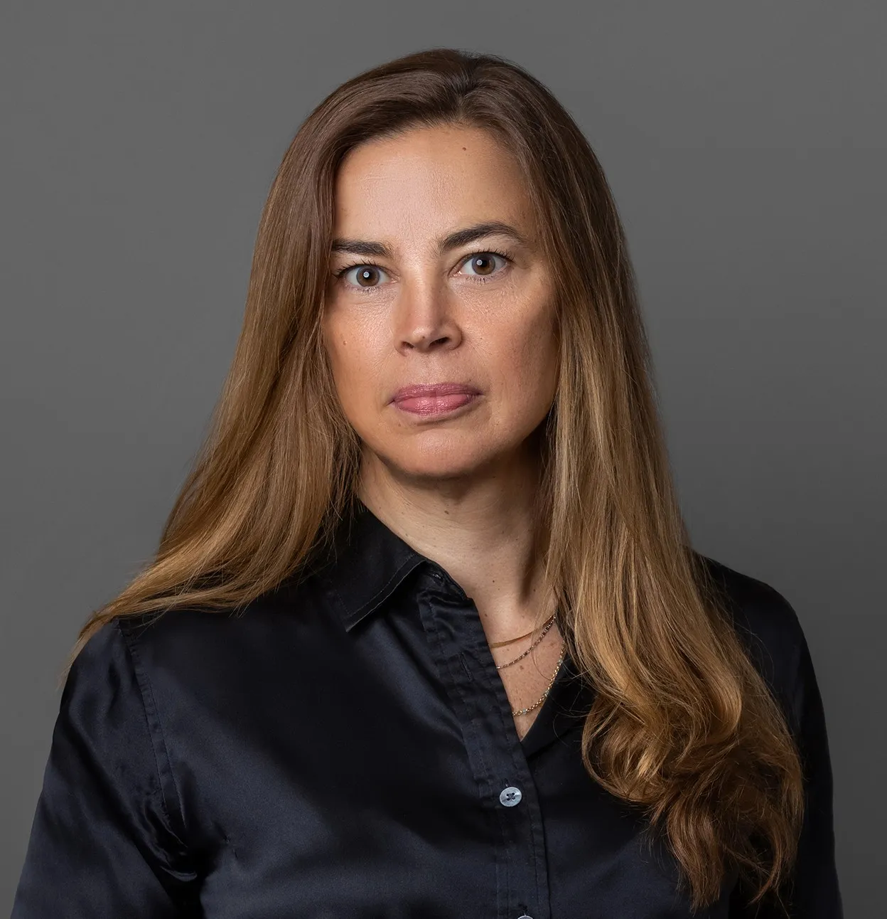 Portrait of a woman with long light brown hair wearing a black collared shirt against a plain gray background.