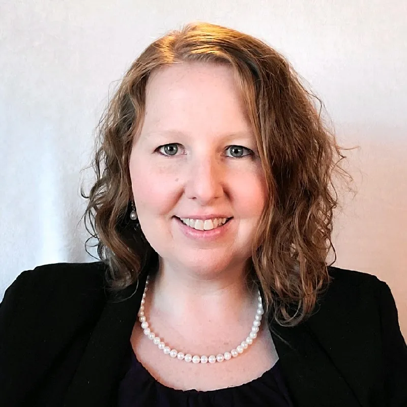 Smiling woman with shoulder-length curly hair wearing a black blazer and pearl necklace.