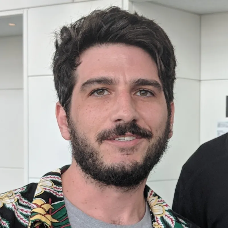 Man with dark hair and beard wearing a patterned shirt and gray t-shirt, smiling slightly indoors.