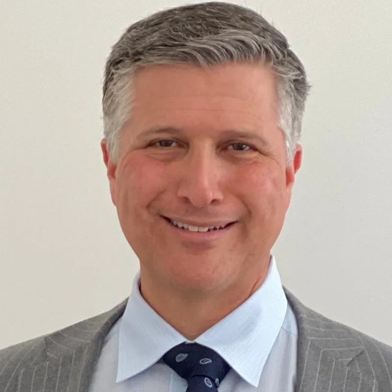Man with short gray hair wearing a striped gray suit, light blue dress shirt, and dark blue paisley tie, smiling in front of a plain background.