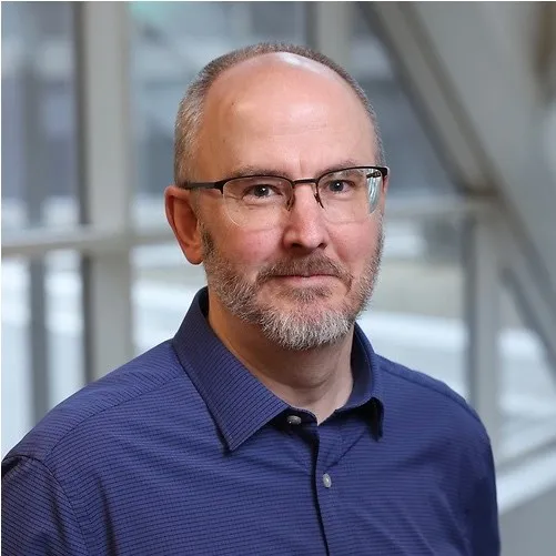 Middle-aged man with glasses, short gray beard, and blue collared shirt, standing indoors with blurred window background.