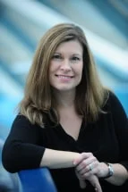 Smiling woman with shoulder-length brown hair wearing a black top, seated with arms crossed on a blue surface.