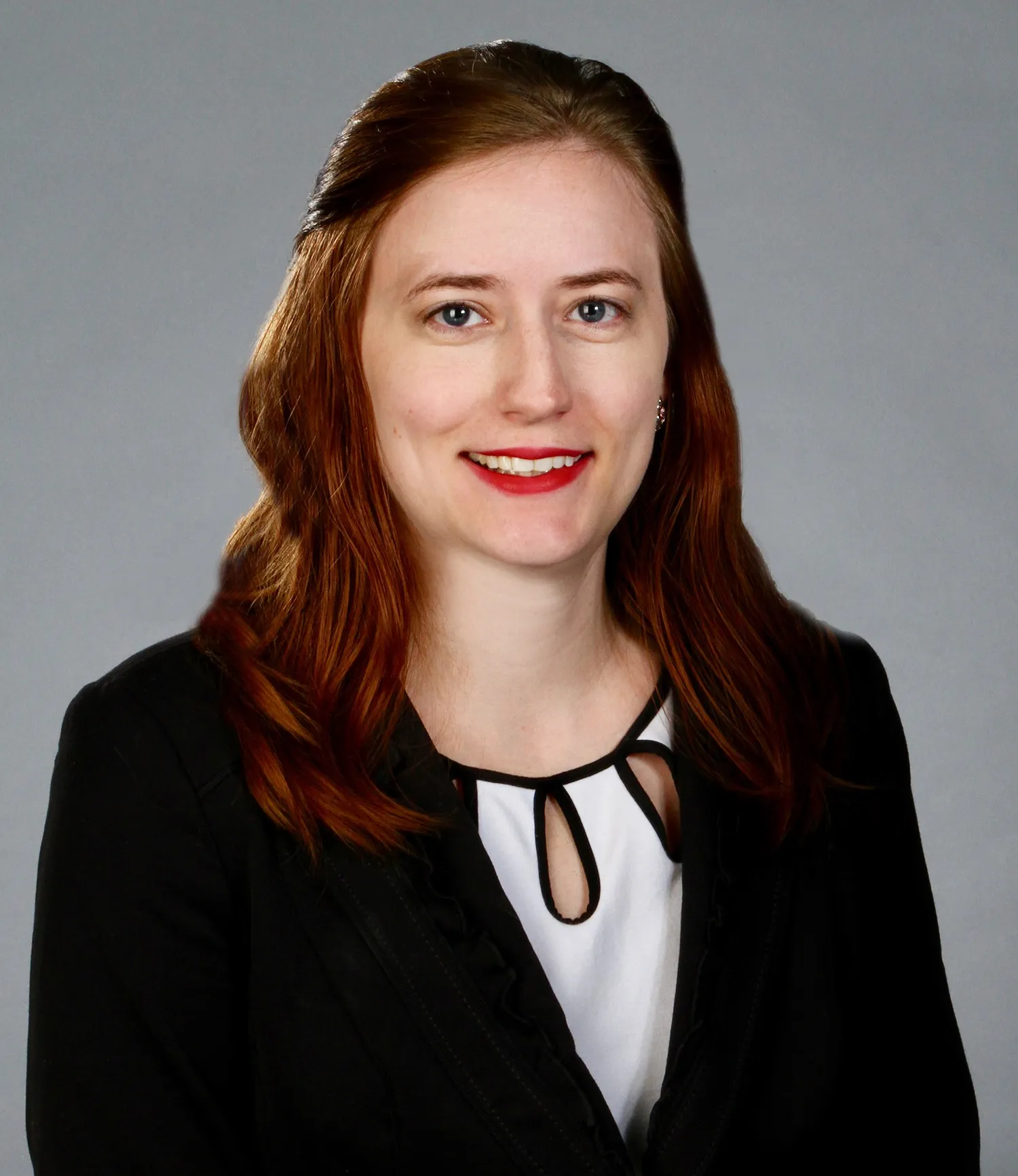 Smiling woman with red hair wearing a black blazer and white blouse with black trim against a gray background.