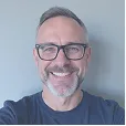 Smiling middle-aged man with glasses and short gray hair wearing a dark shirt against a plain light background.