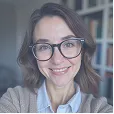 Smiling woman with glasses and shoulder-length brown hair in an indoor setting with bookshelves in the background.