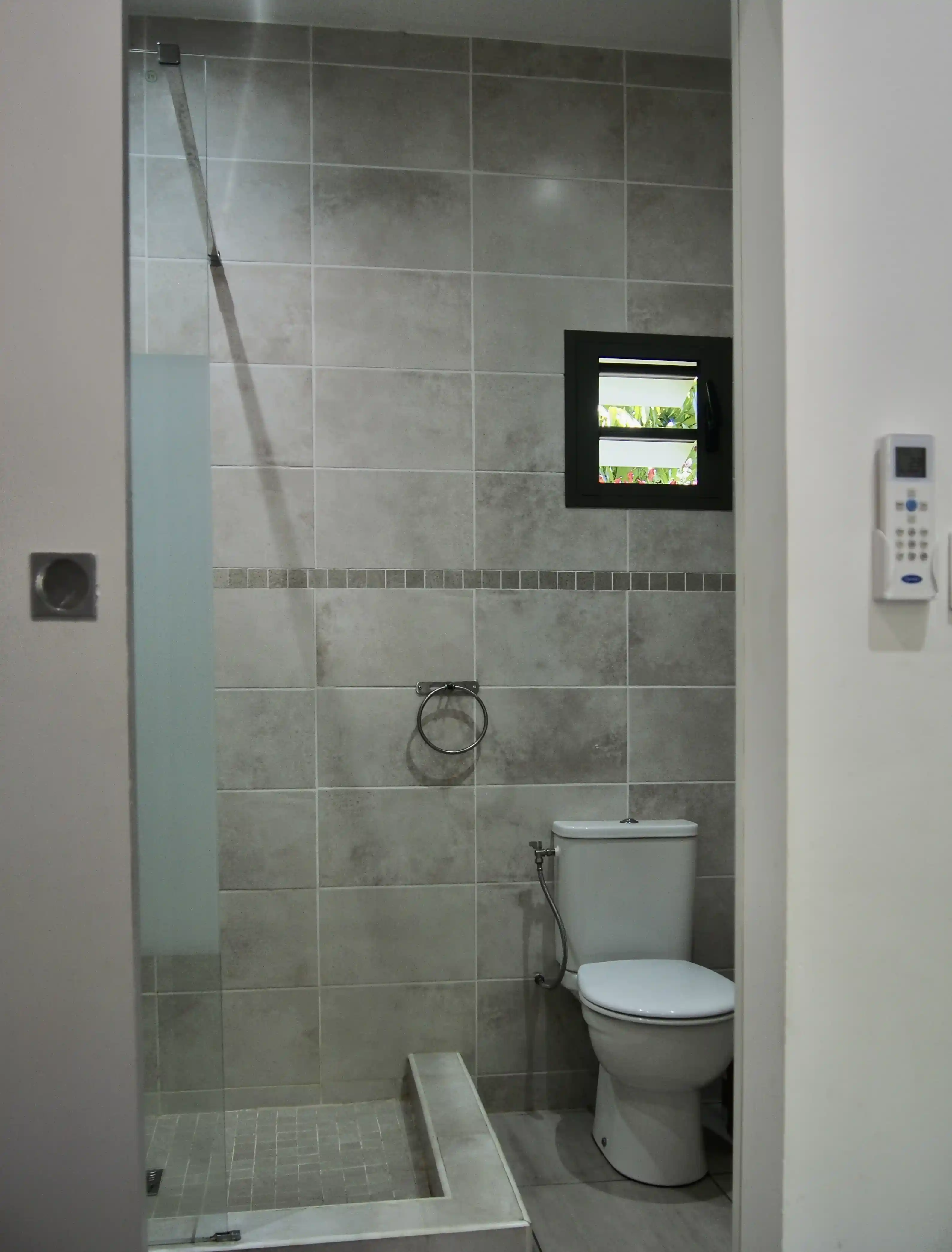 Modern bathroom with gray tiled walls, a white toilet, a towel ring, and a walk-in shower with a glass partition.