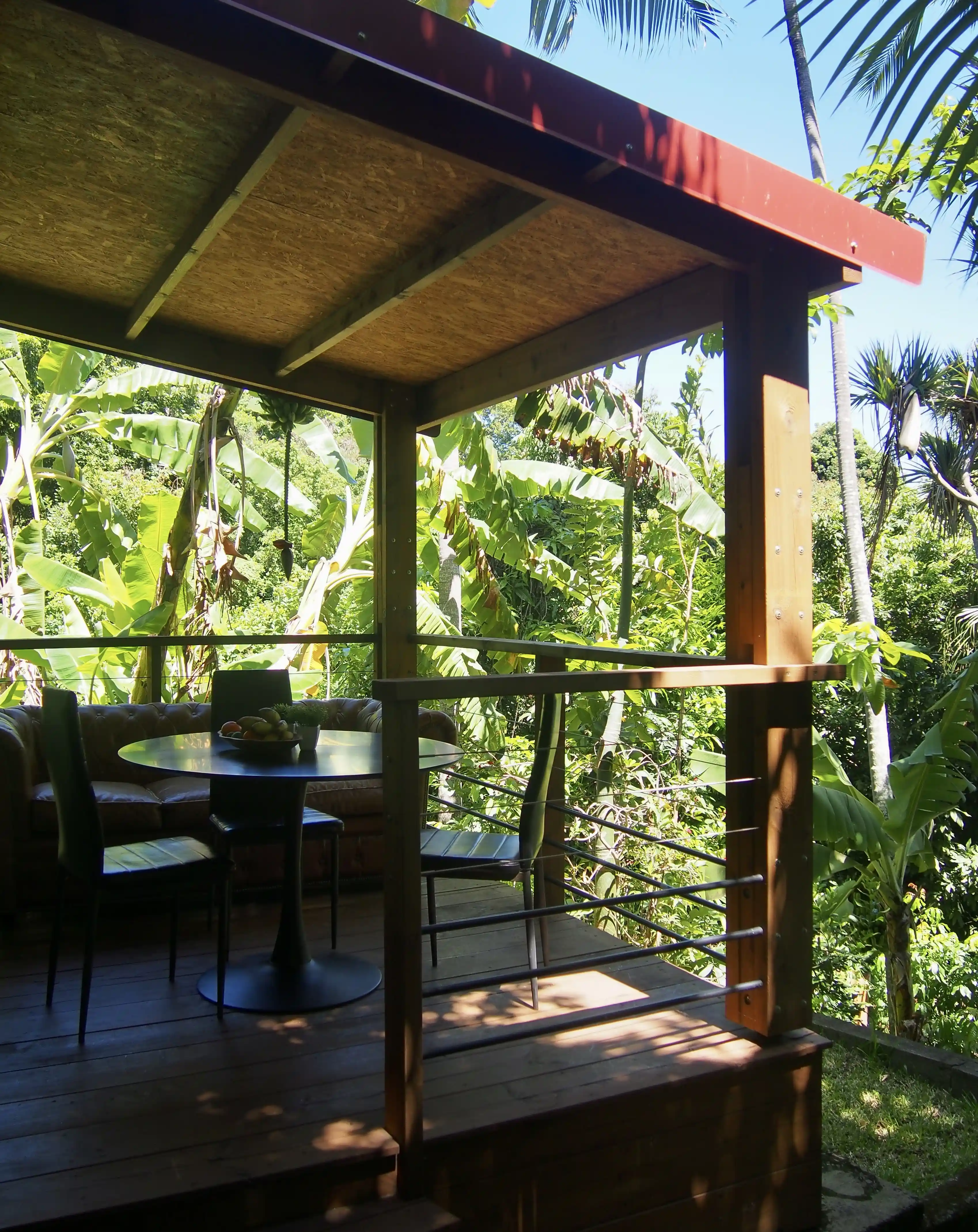 Covered wooden patio with a round table, chairs, and a sofa surrounded by lush tropical greenery.