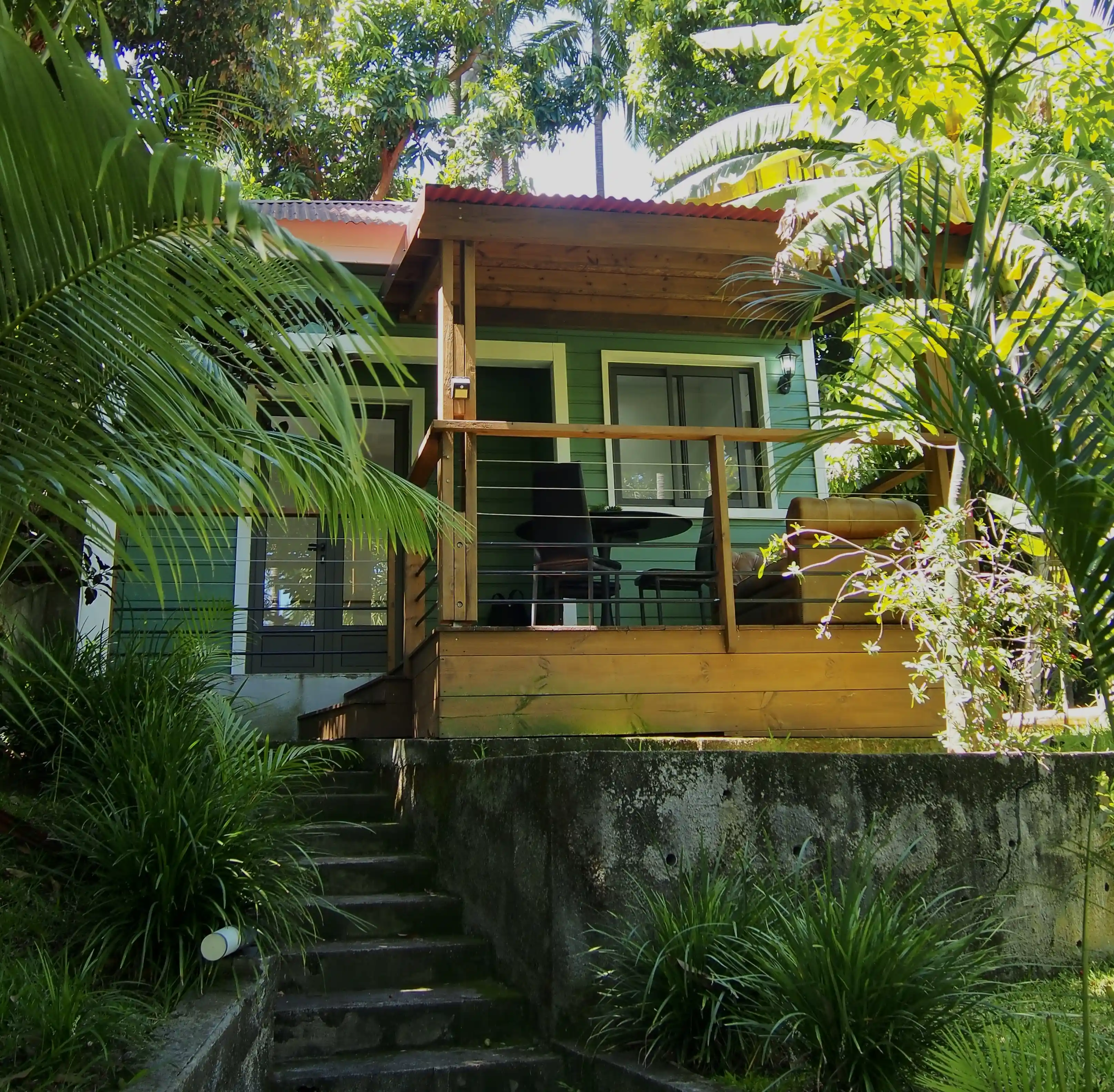 Small green cabin with a wooden porch, outdoor table, chairs, and surrounded by lush tropical plants.