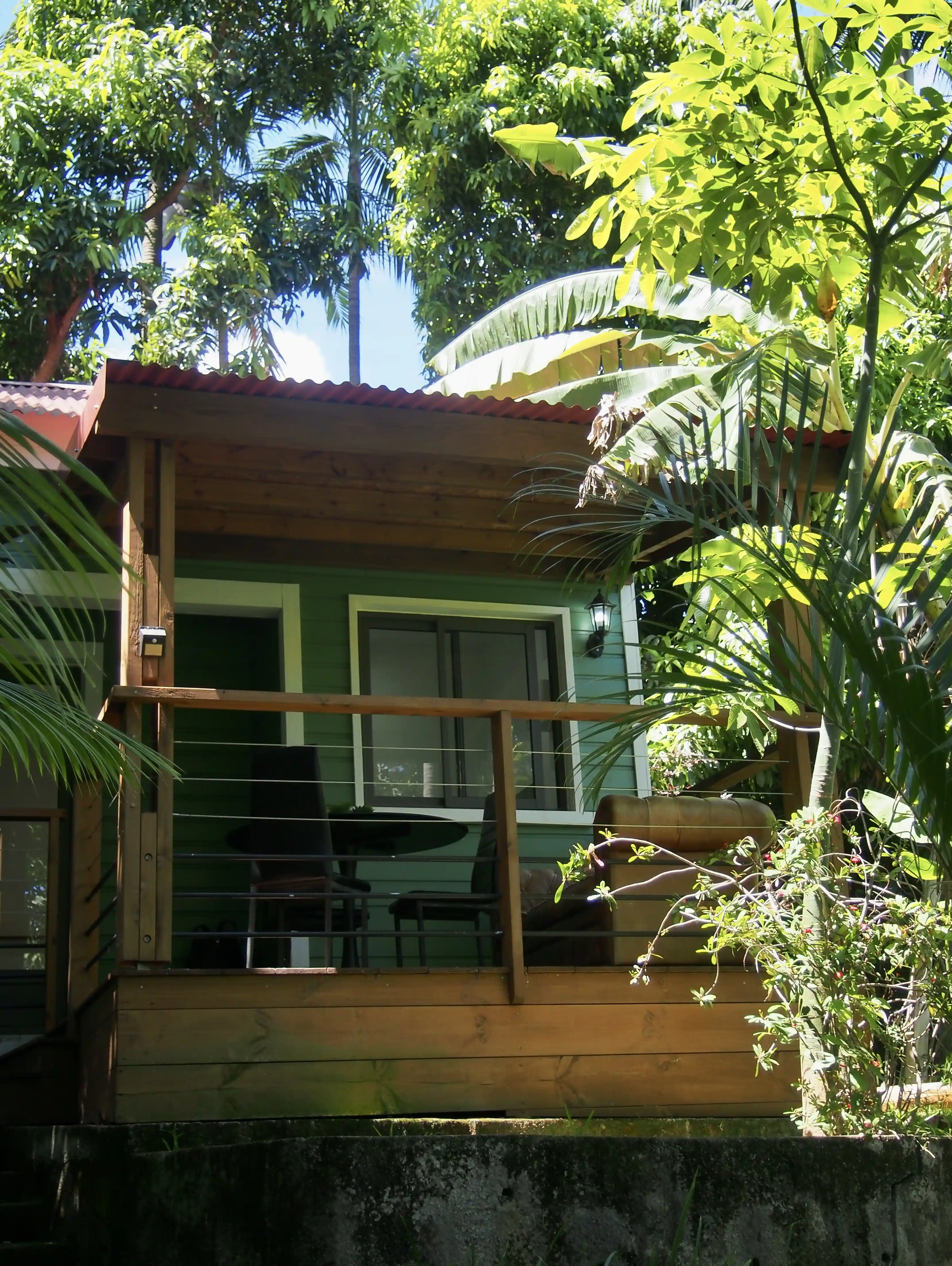 Wooden porch with table, chairs, and a sofa surrounded by lush tropical greenery and trees.
