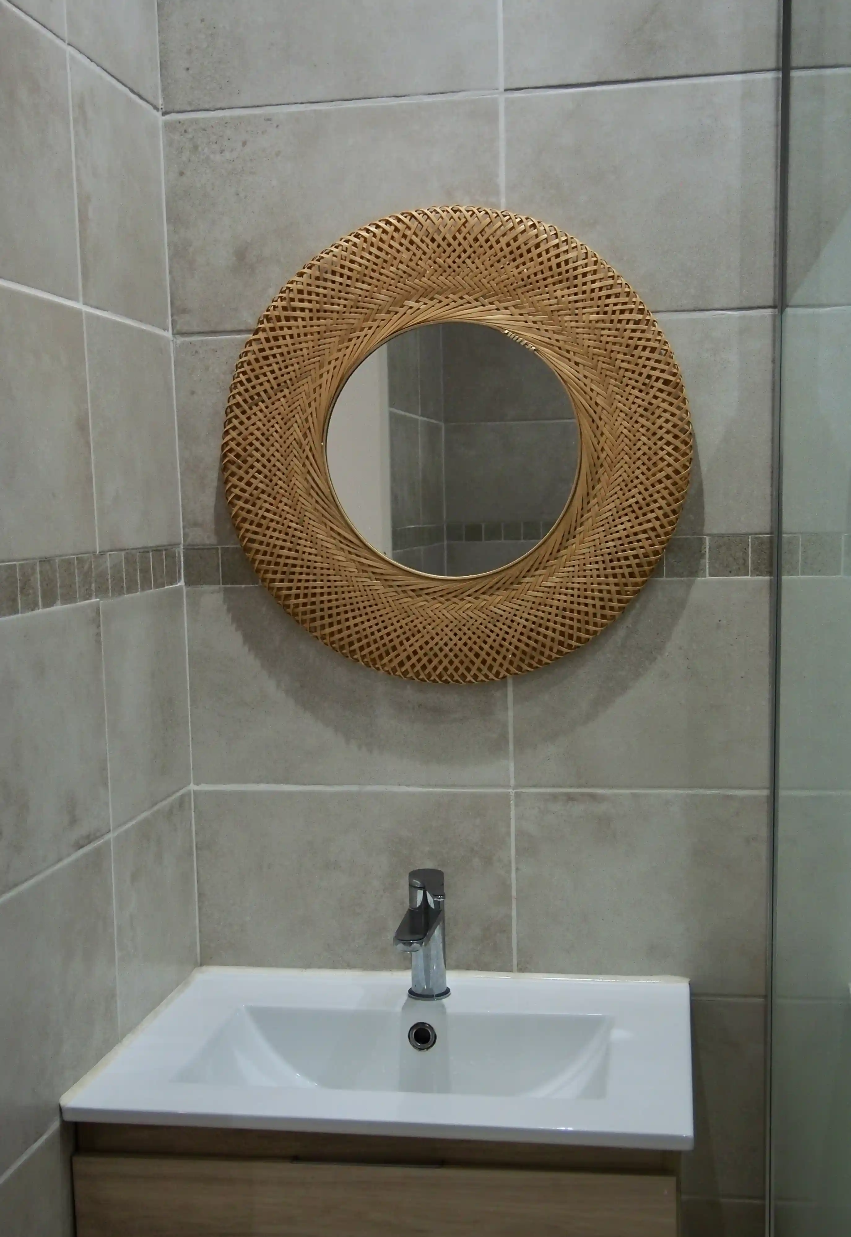 Modern bathroom sink with a chrome faucet below a round woven wicker-framed mirror on tiled walls.