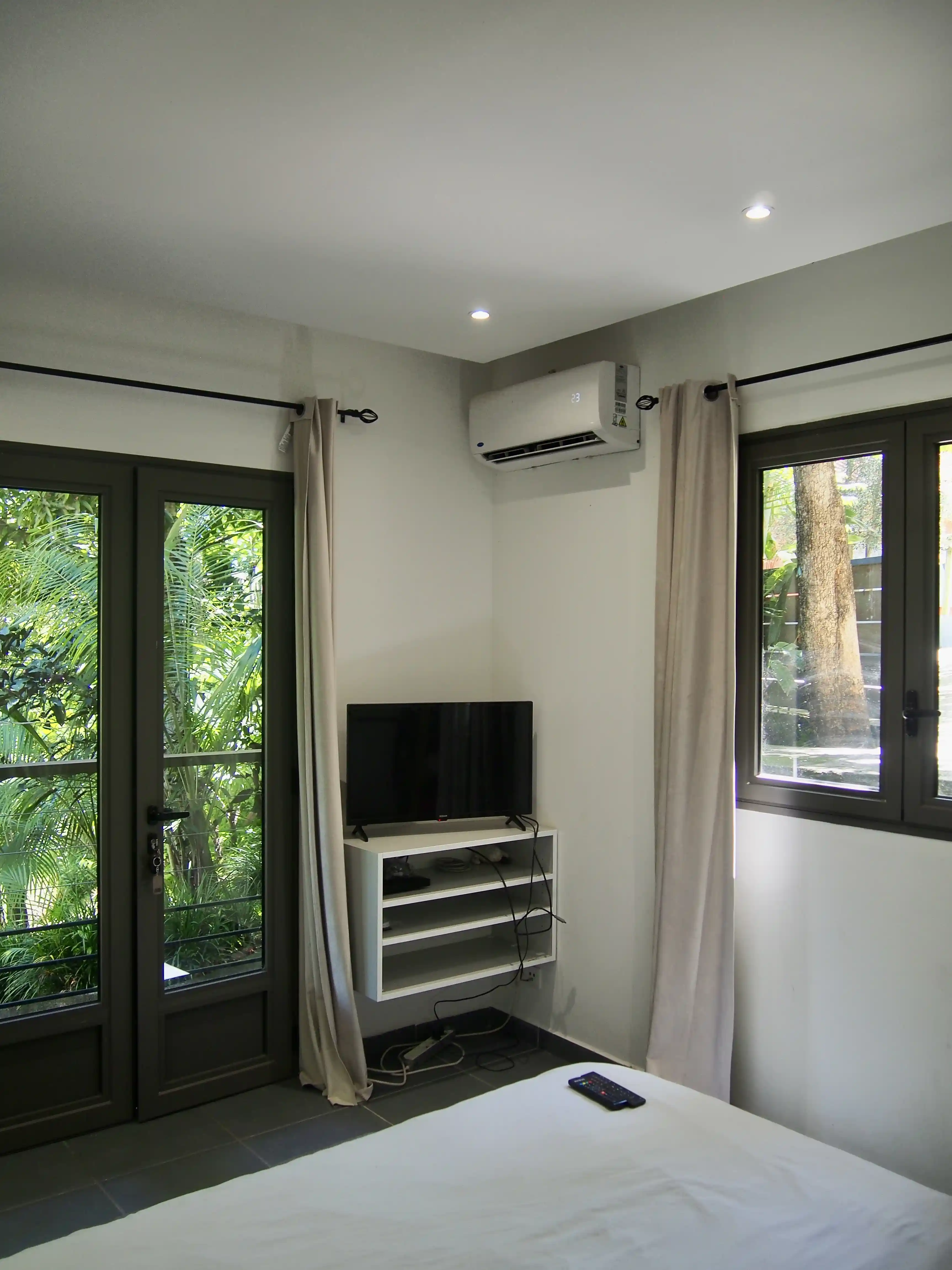 Corner of a bedroom with white walls, a mounted air conditioner, a TV on a floating shelf, beige curtains, a bed with white bedding, and glass doors and windows showing green outdoor foliage.
