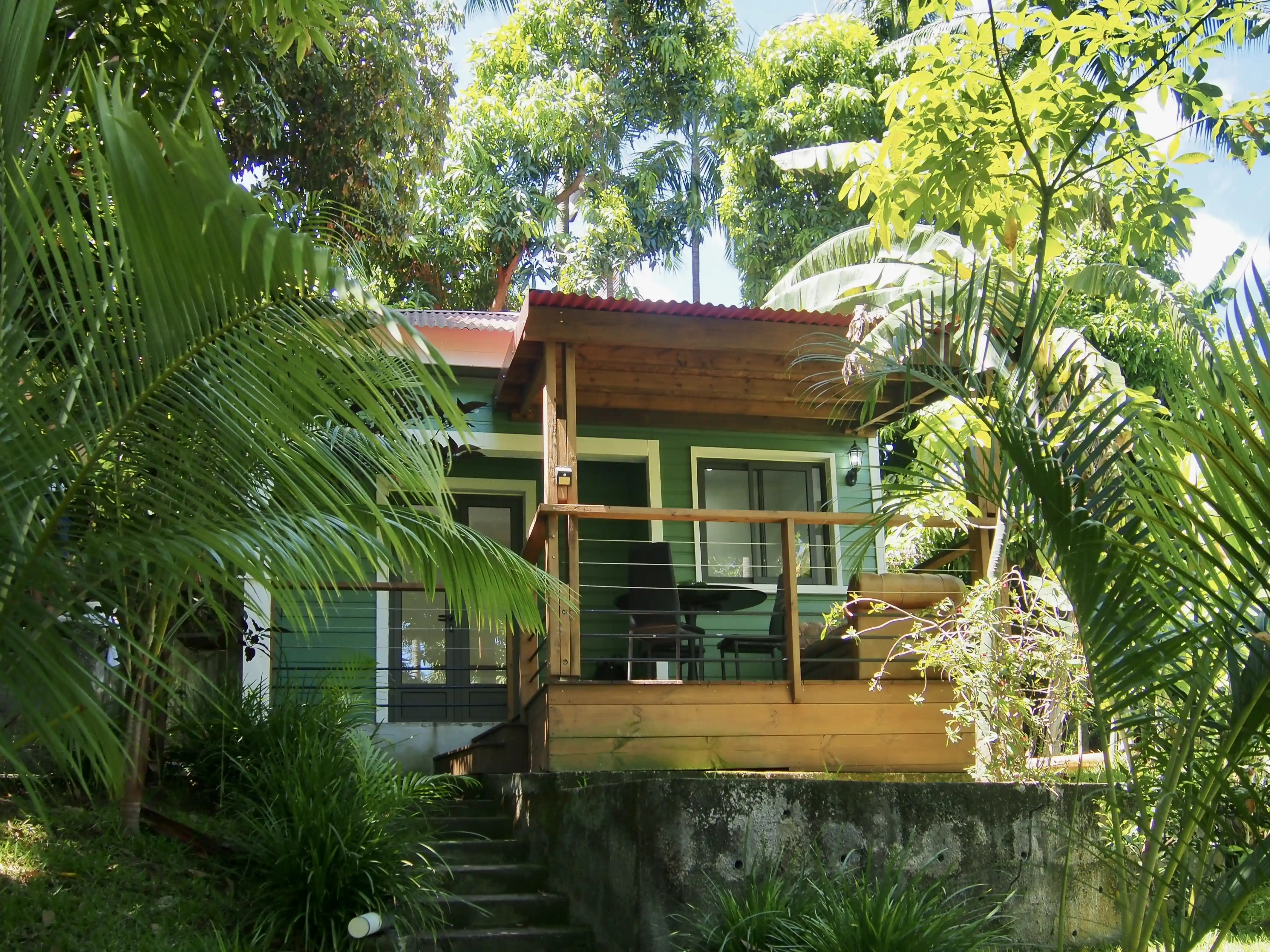 Small green house with wooden porch surrounded by lush tropical plants and trees.