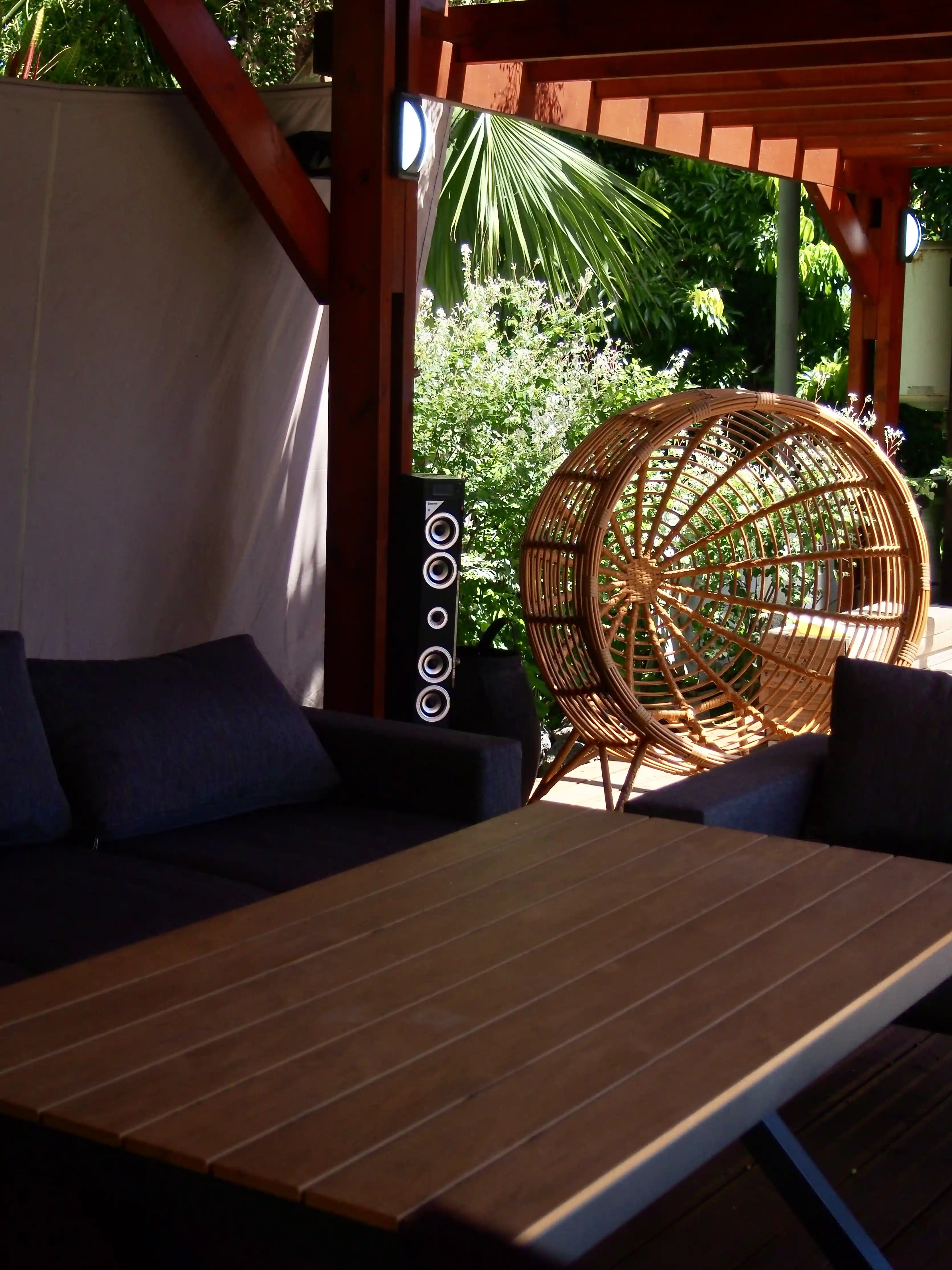 Outdoor seating area with dark cushioned sofas, a wooden table, a round rattan chair, and green foliage under a wooden pergola.