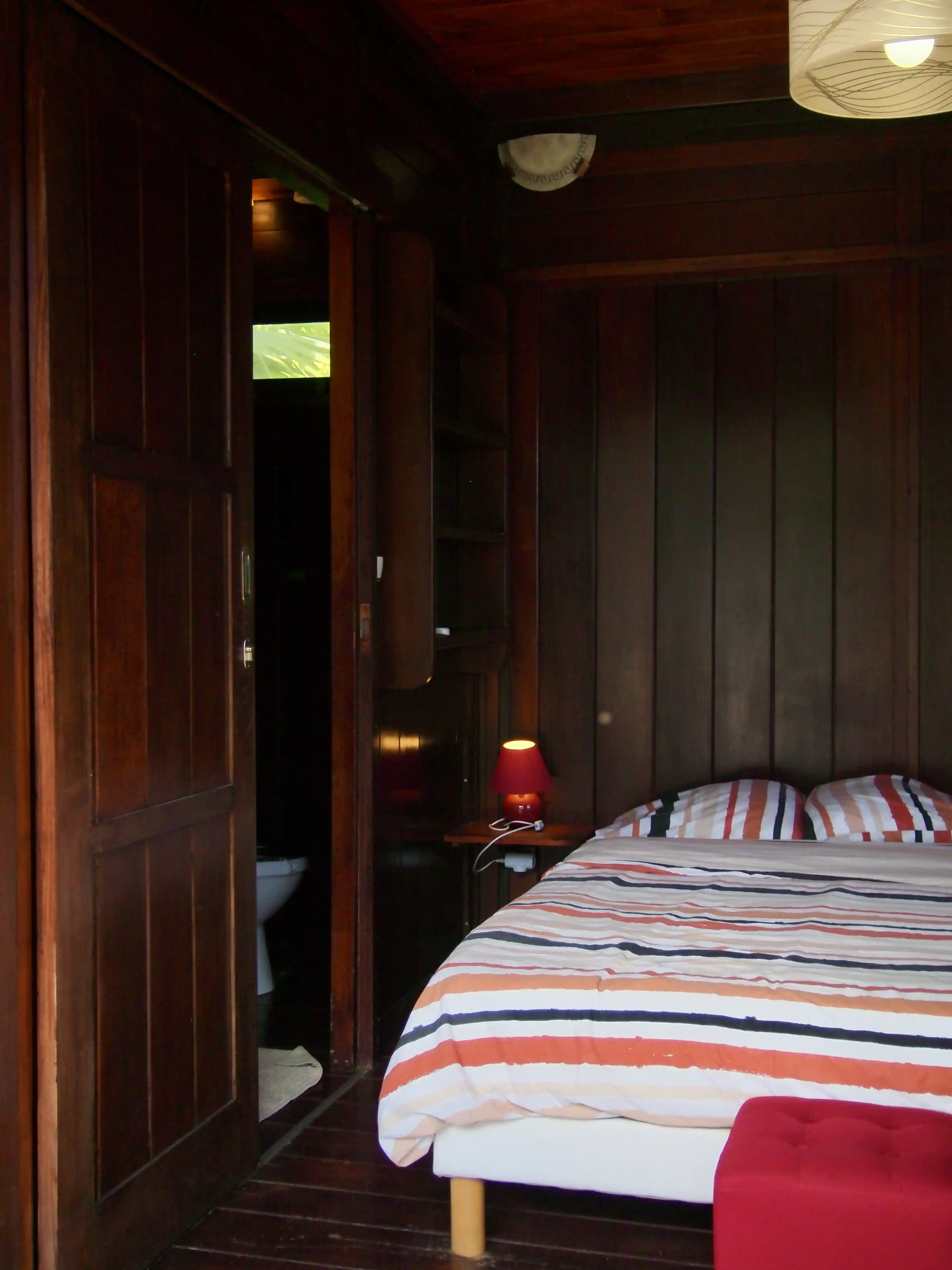 Cozy bedroom with wooden walls, striped bedding, a red bedside lamp, and an open door leading to a bathroom.