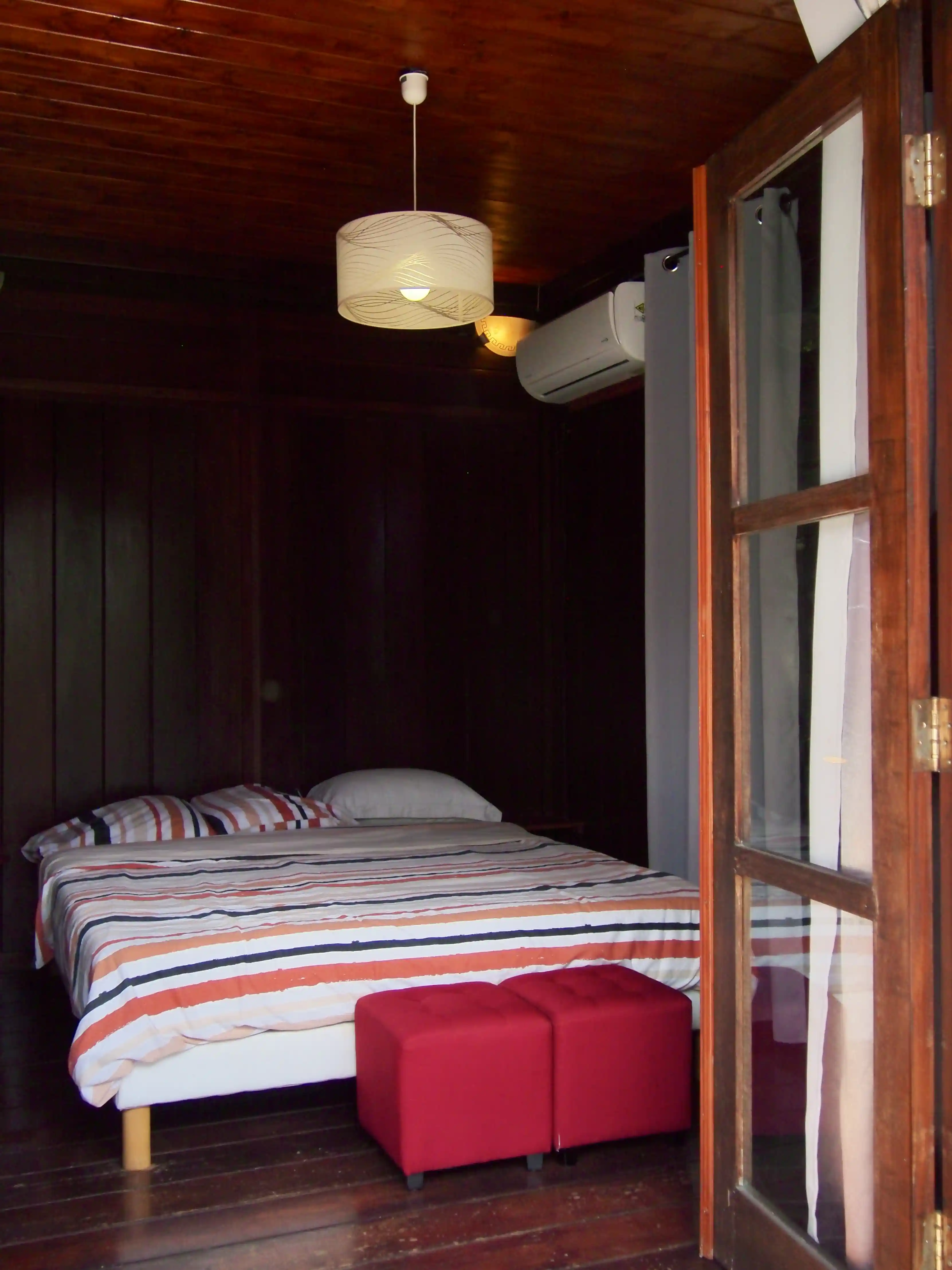 Cozy bedroom with wooden walls and ceiling, striped bedding on bed, two red ottomans, and an air conditioner.