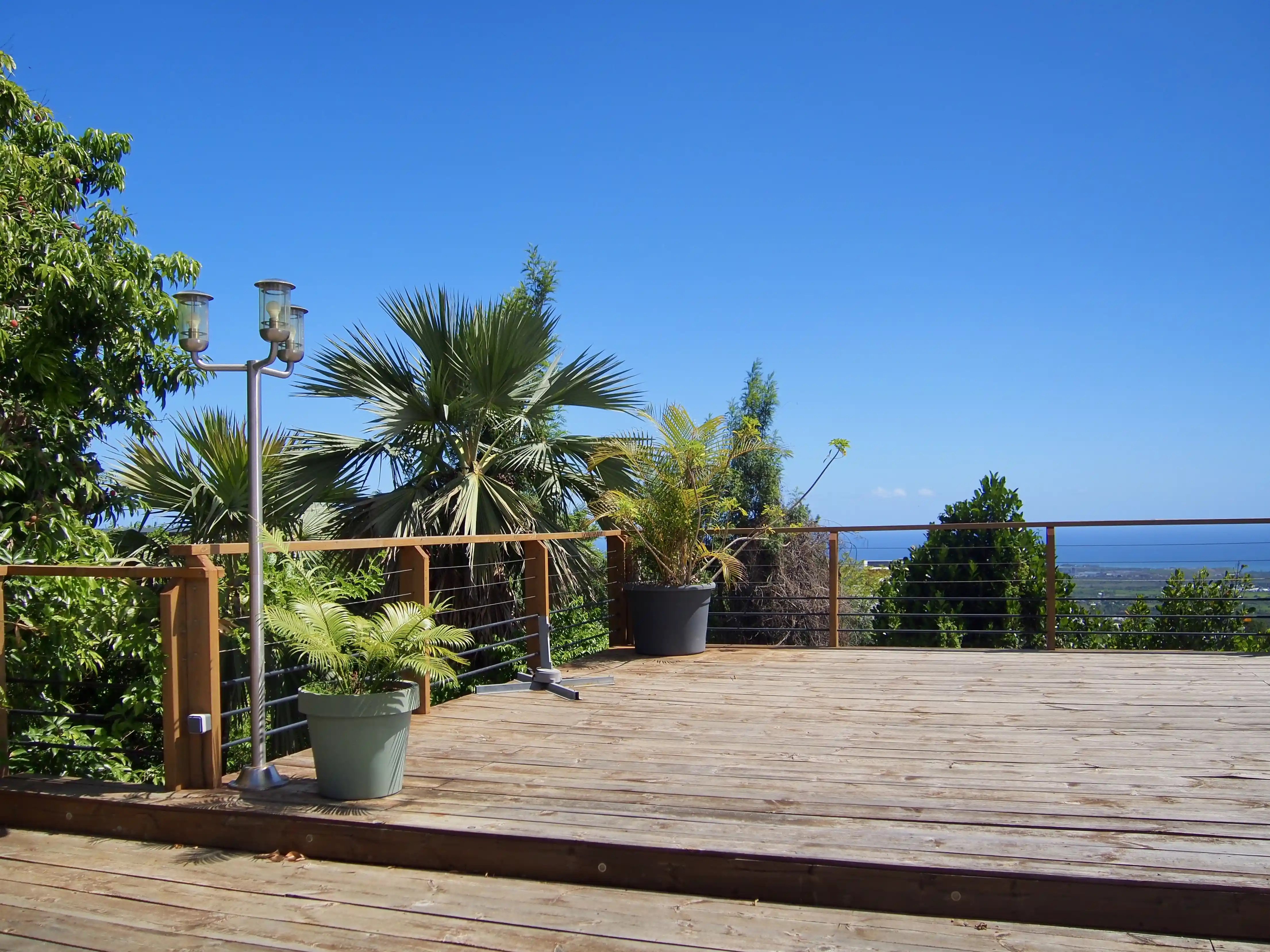 Spacious wooden deck with potted plants and a metal lamp post overlooking green trees and a distant ocean under a clear blue sky.