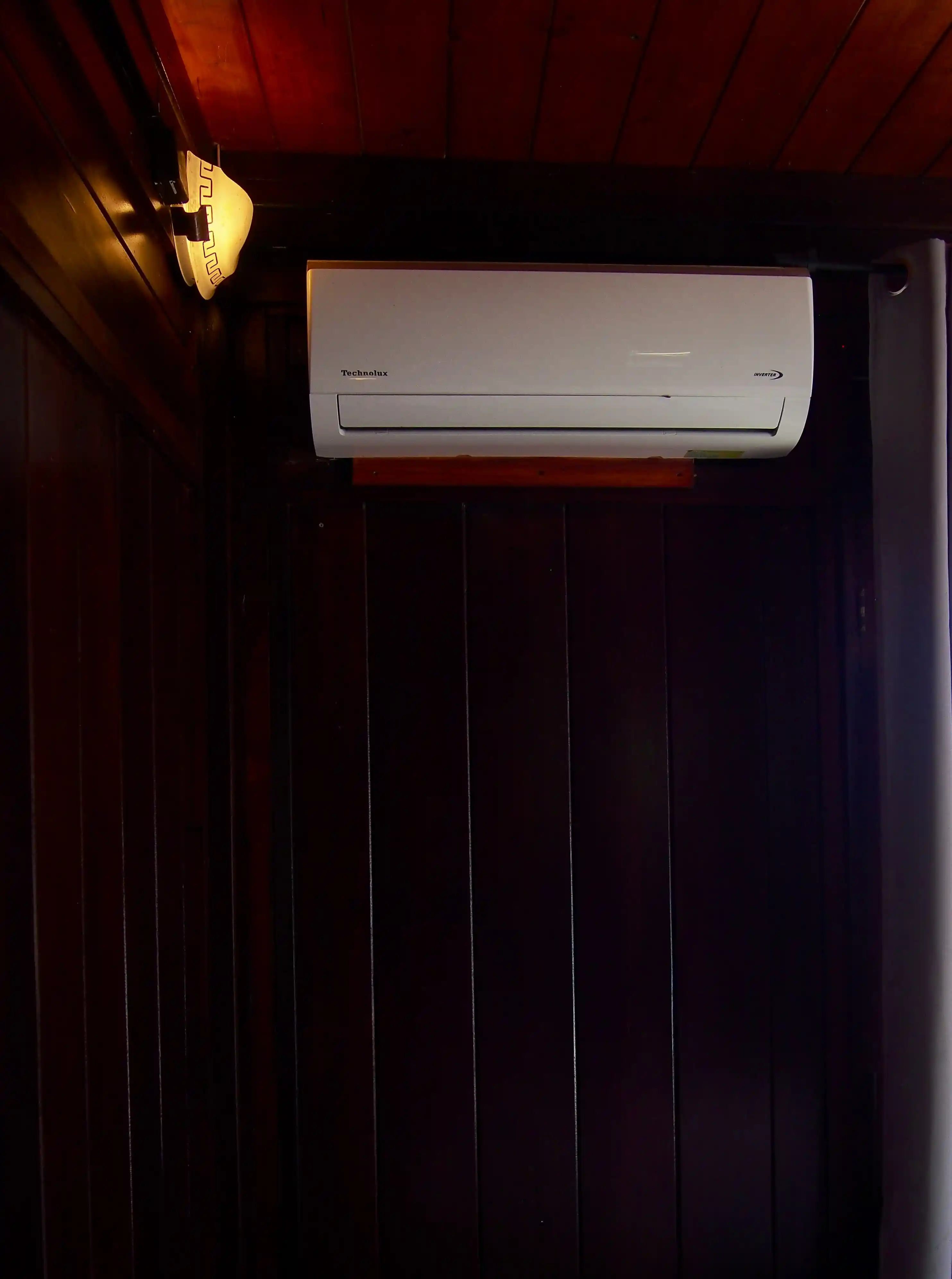 White Technolux wall-mounted air conditioner on dark wooden panel wall with a lit vintage-style wall lamp to the left.