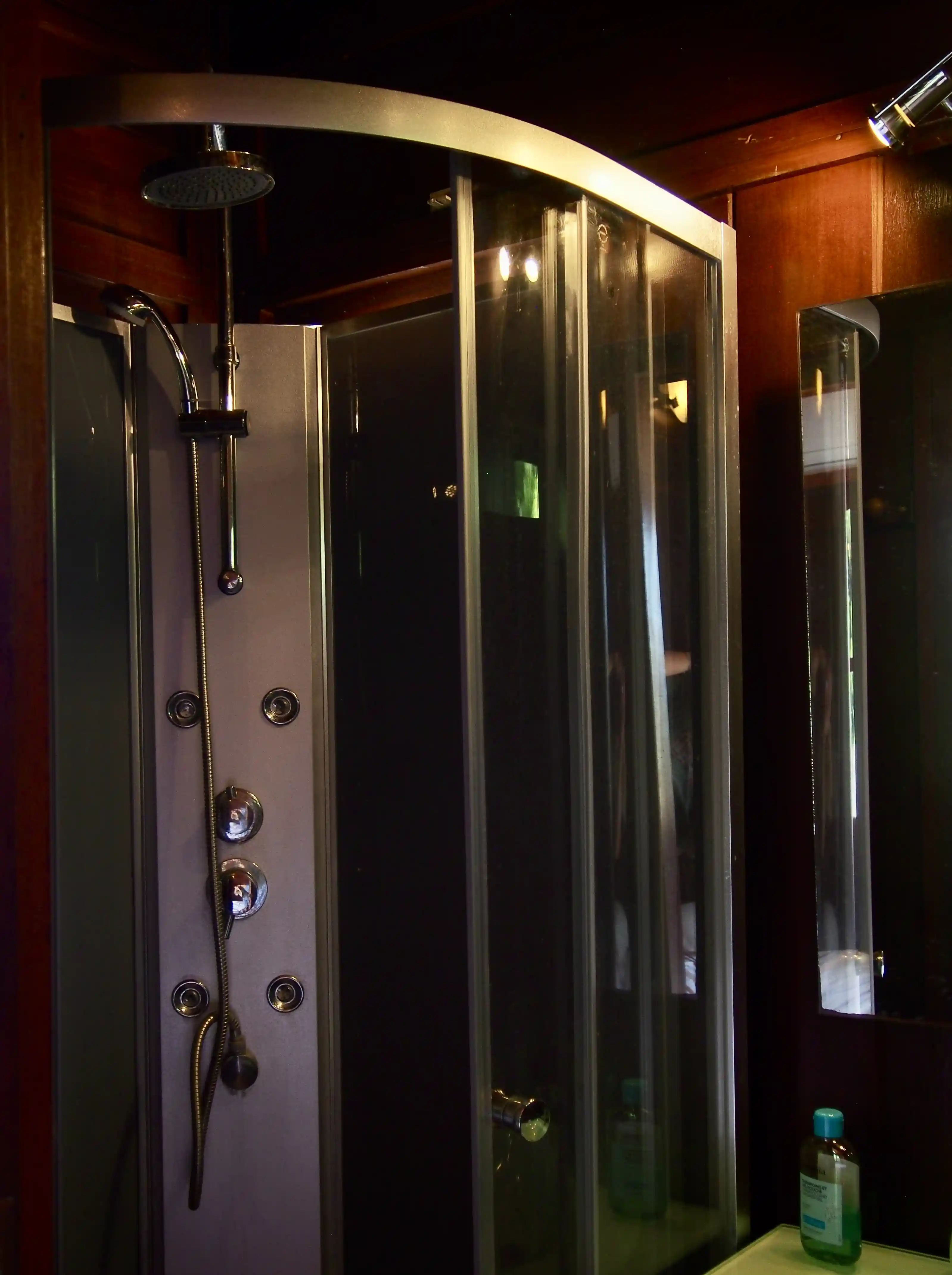 Modern shower cabin with glass doors, adjustable showerhead, and a bottle of shampoo on a nearby shelf.