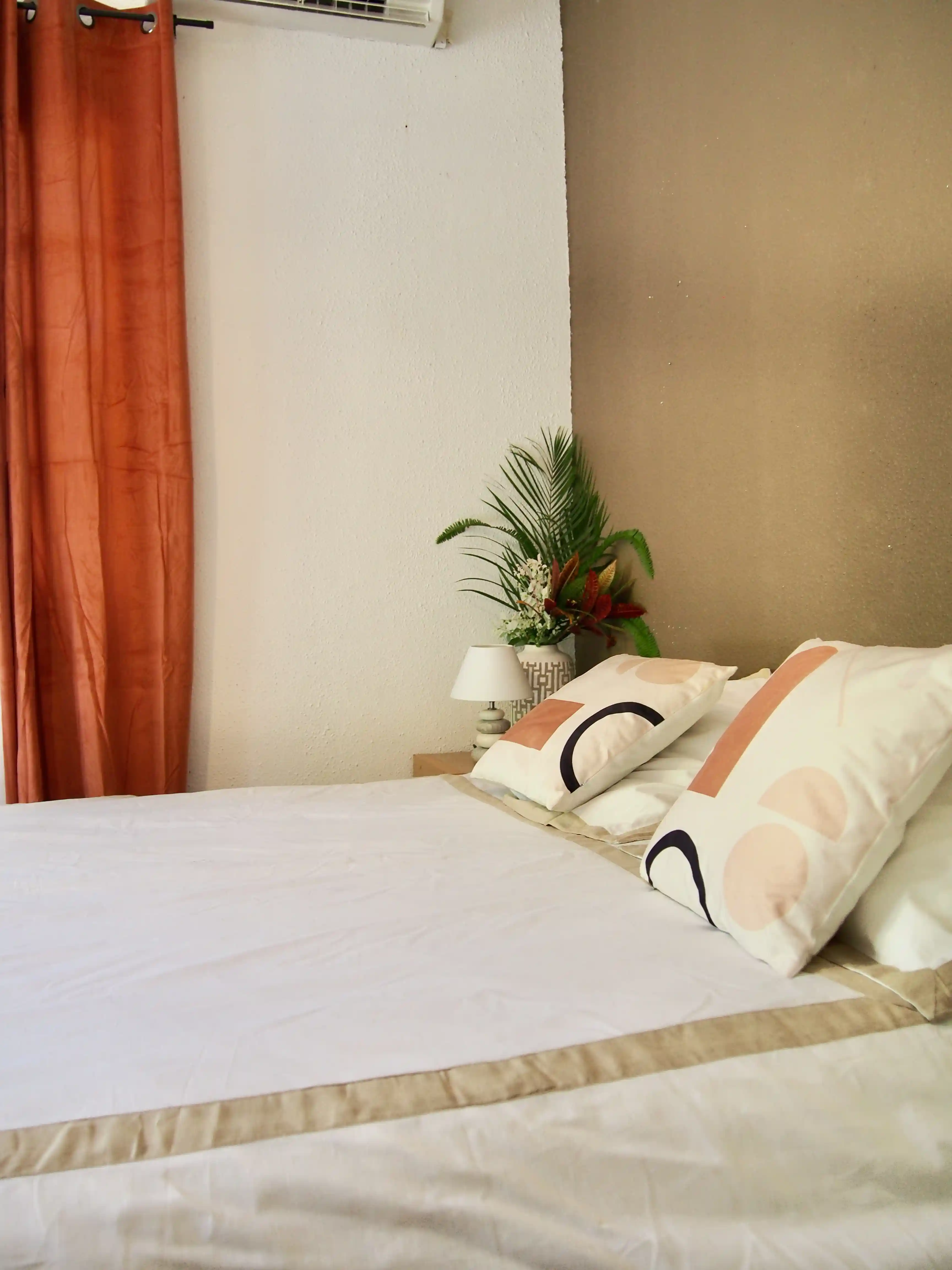 Neatly made bed with white and beige bedding, decorative pillows, a bedside lamp, and a plant in a vase next to a brown wall and orange curtain.