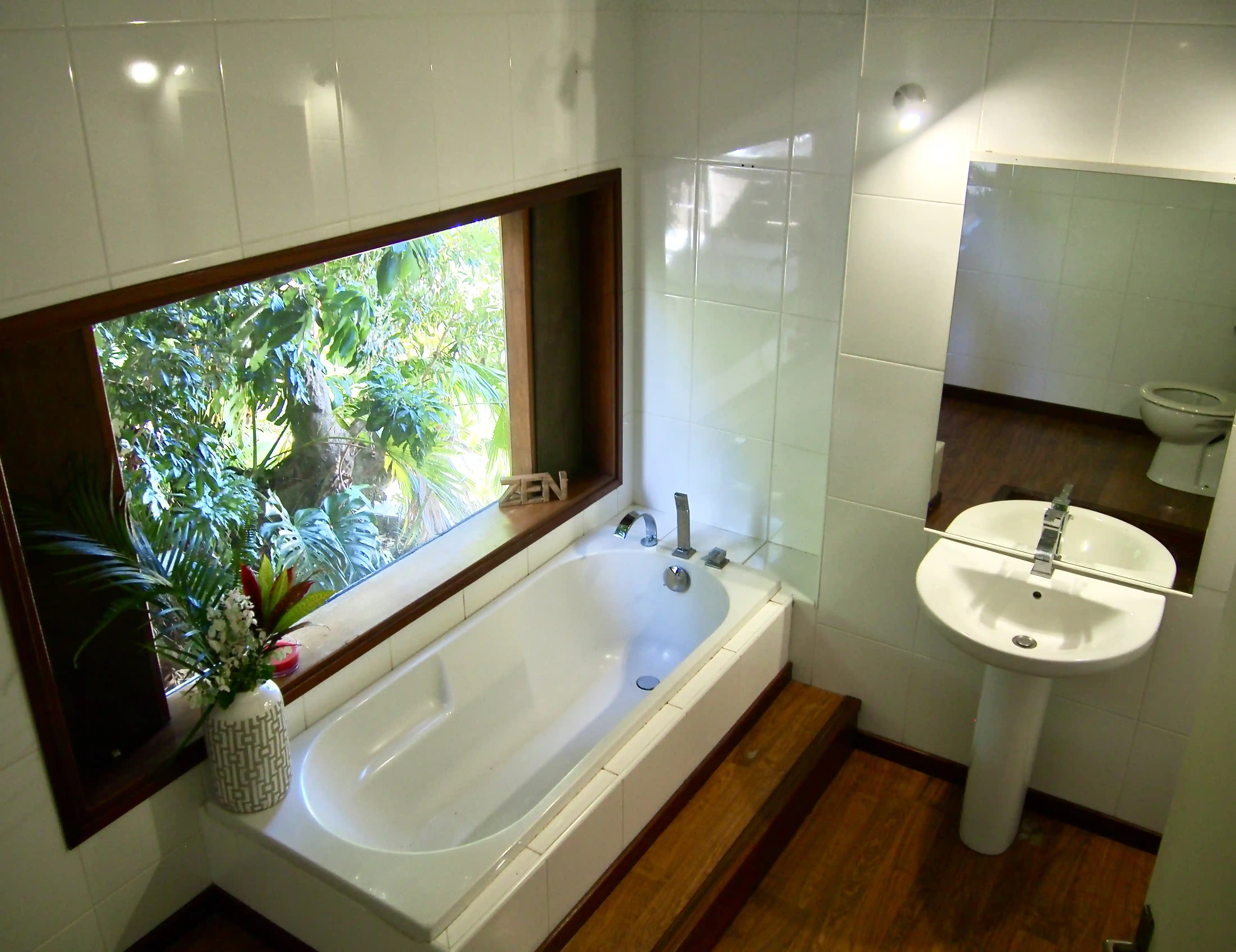 Bathroom with a white bathtub beside a large window showing green foliage, a white pedestal sink with a mirror, and wooden flooring.