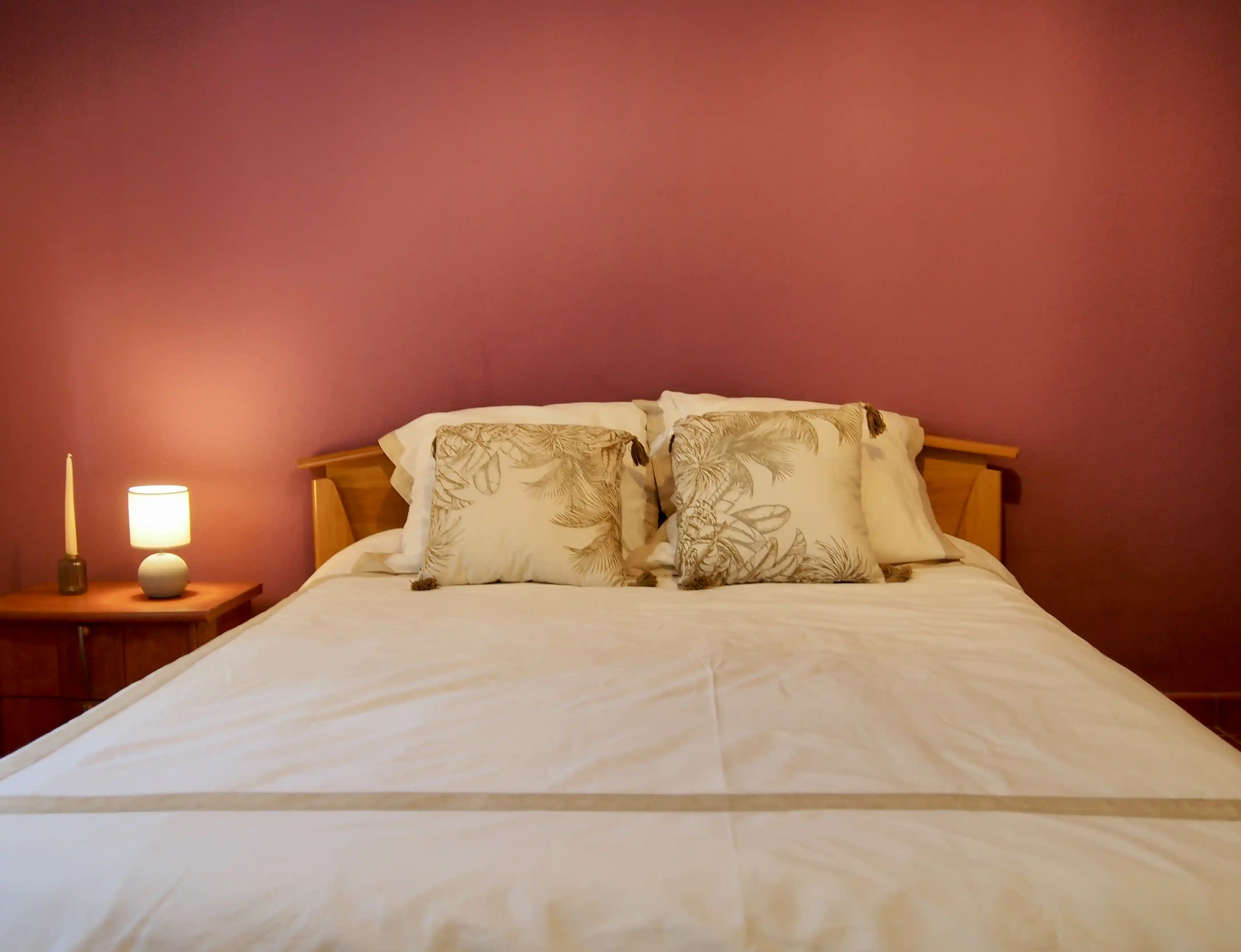 Bed with white linens and decorative beige pillows against a purple wall, with a lit bedside lamp and candle on a wooden nightstand.