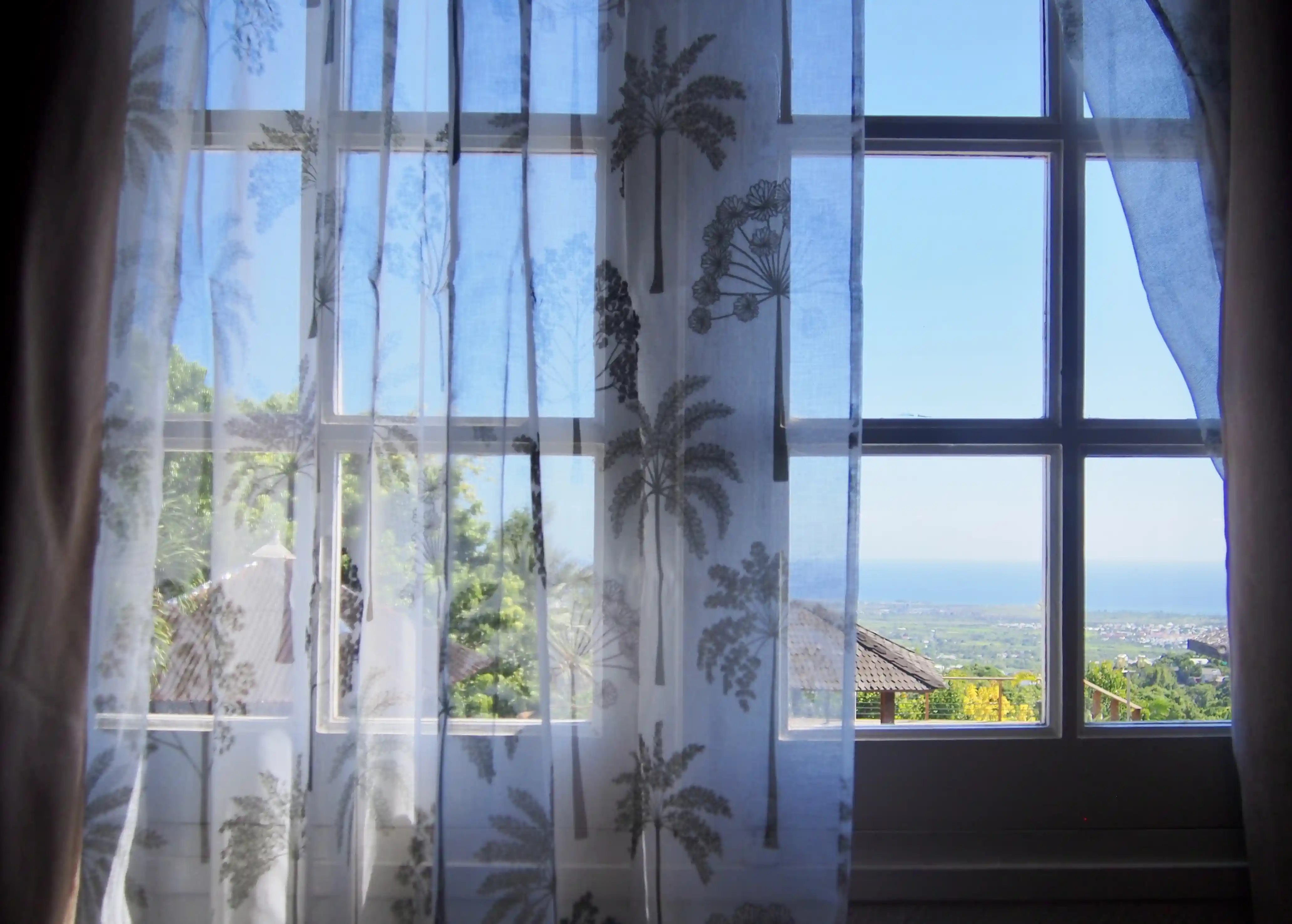 View through a window with sheer curtains patterned with trees, showing a rooftop, green landscape, and distant sea under a clear blue sky.