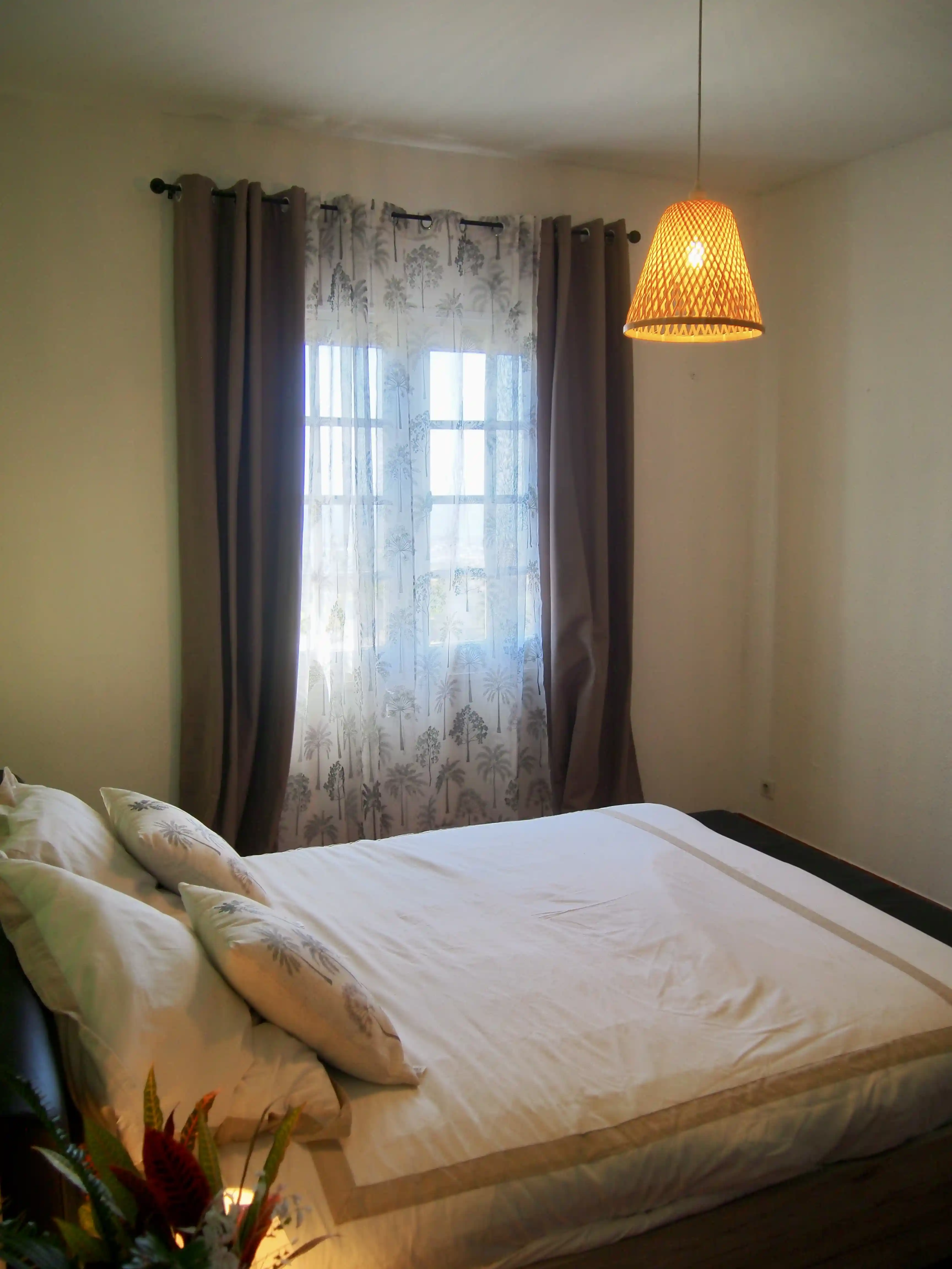 Bed with white linens and patterned pillows next to a window with sheer and dark curtains, illuminated by a hanging lamp.