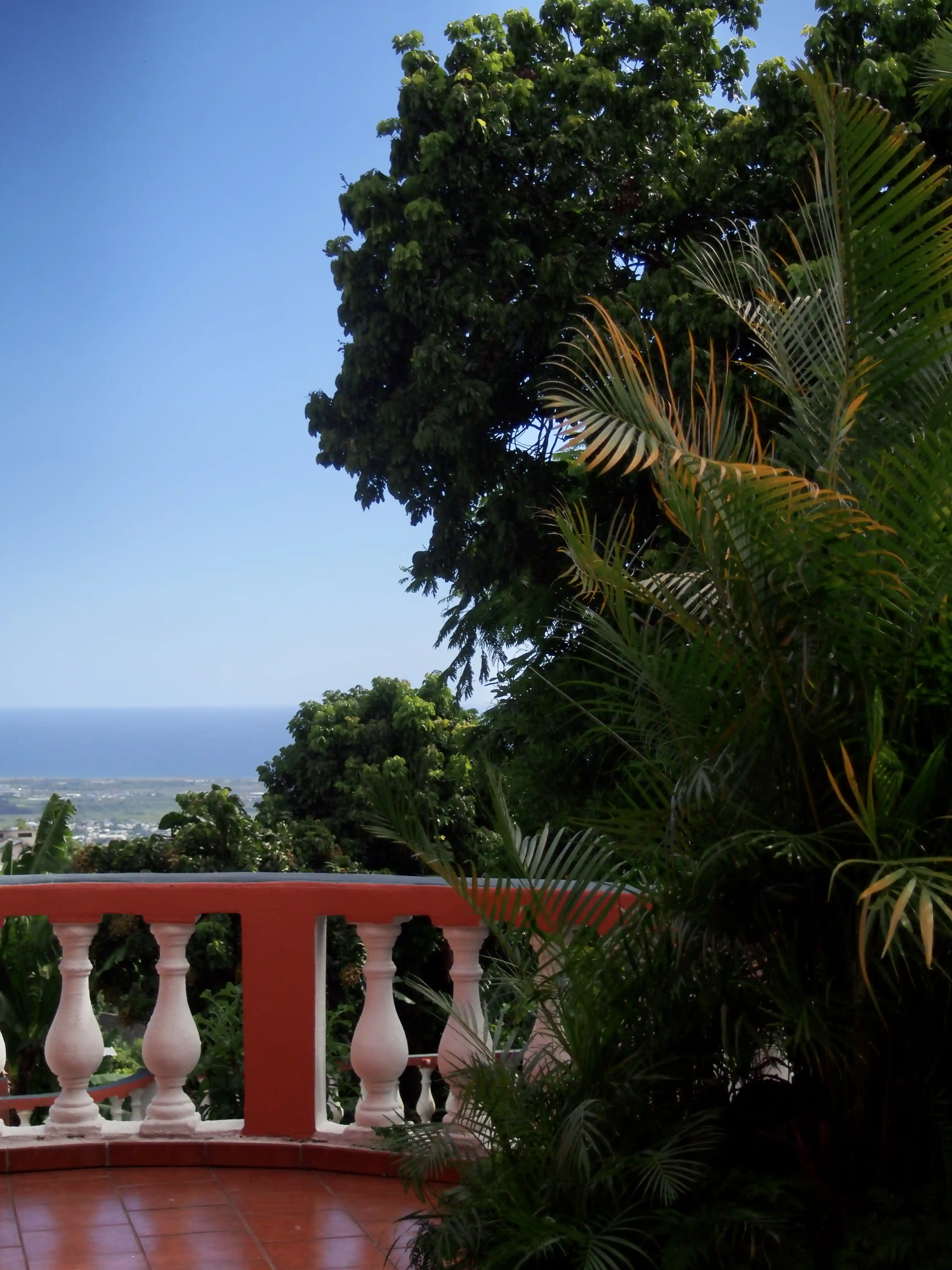 View from a terrace with red-tiled floor and white balustrade overlooking green trees and a distant ocean under a clear blue sky.