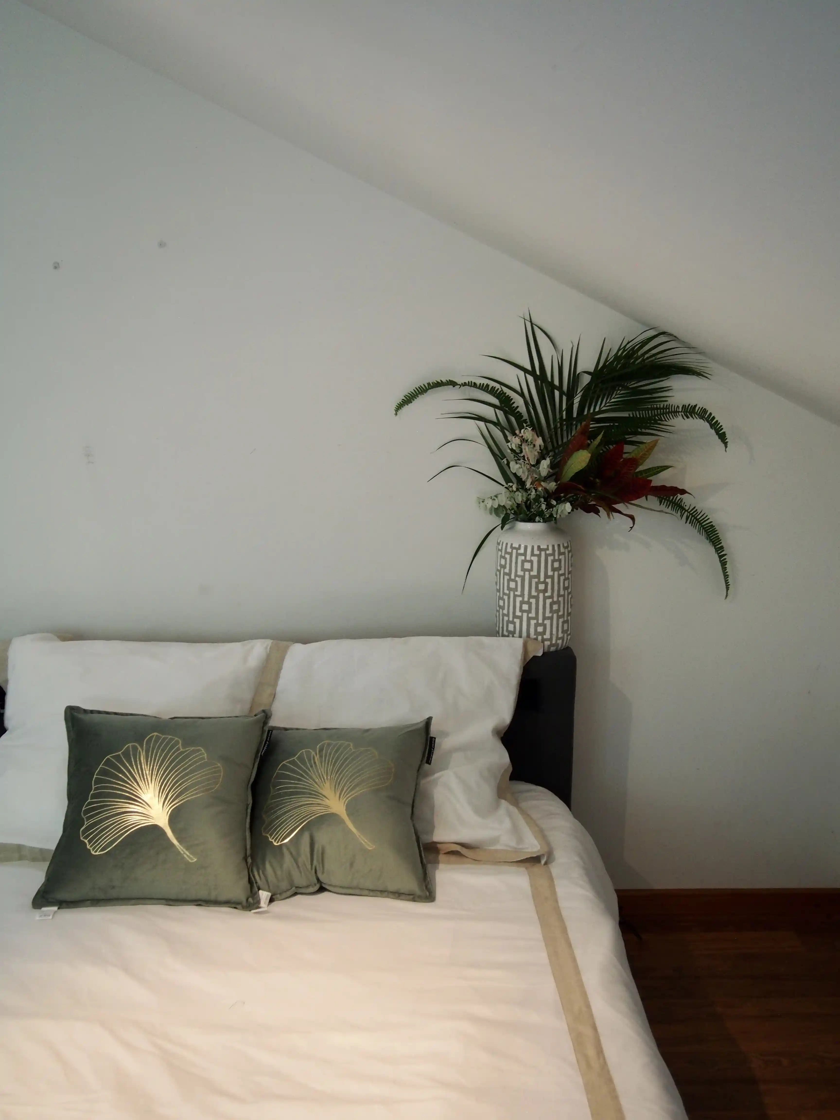Bed with white and beige bedding, two green pillows with gold leaf designs, and a decorative vase with green and red foliage on the headboard.