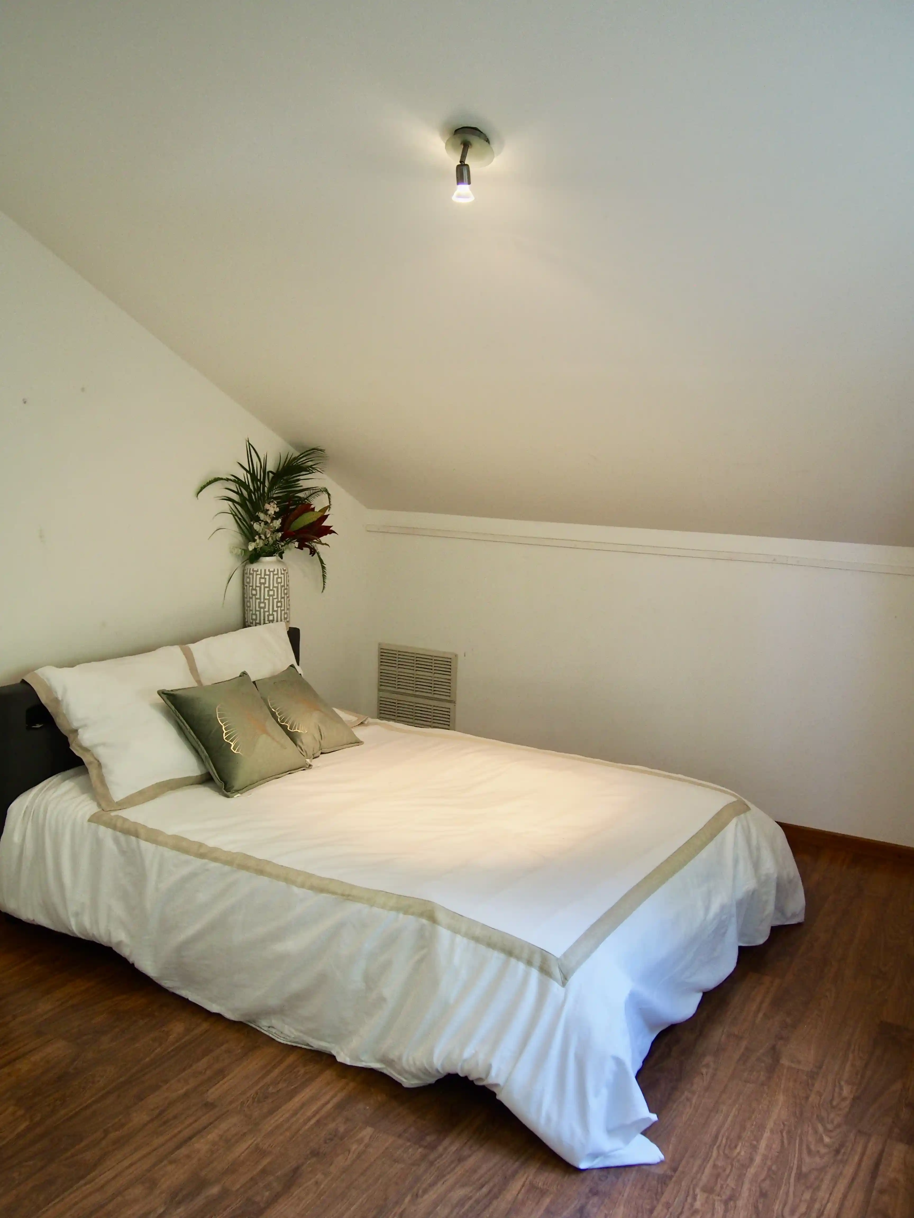 Simple bedroom with a double bed, white and beige bedding, two green decorative pillows, a vase with flowers on the headboard, and a wooden floor.