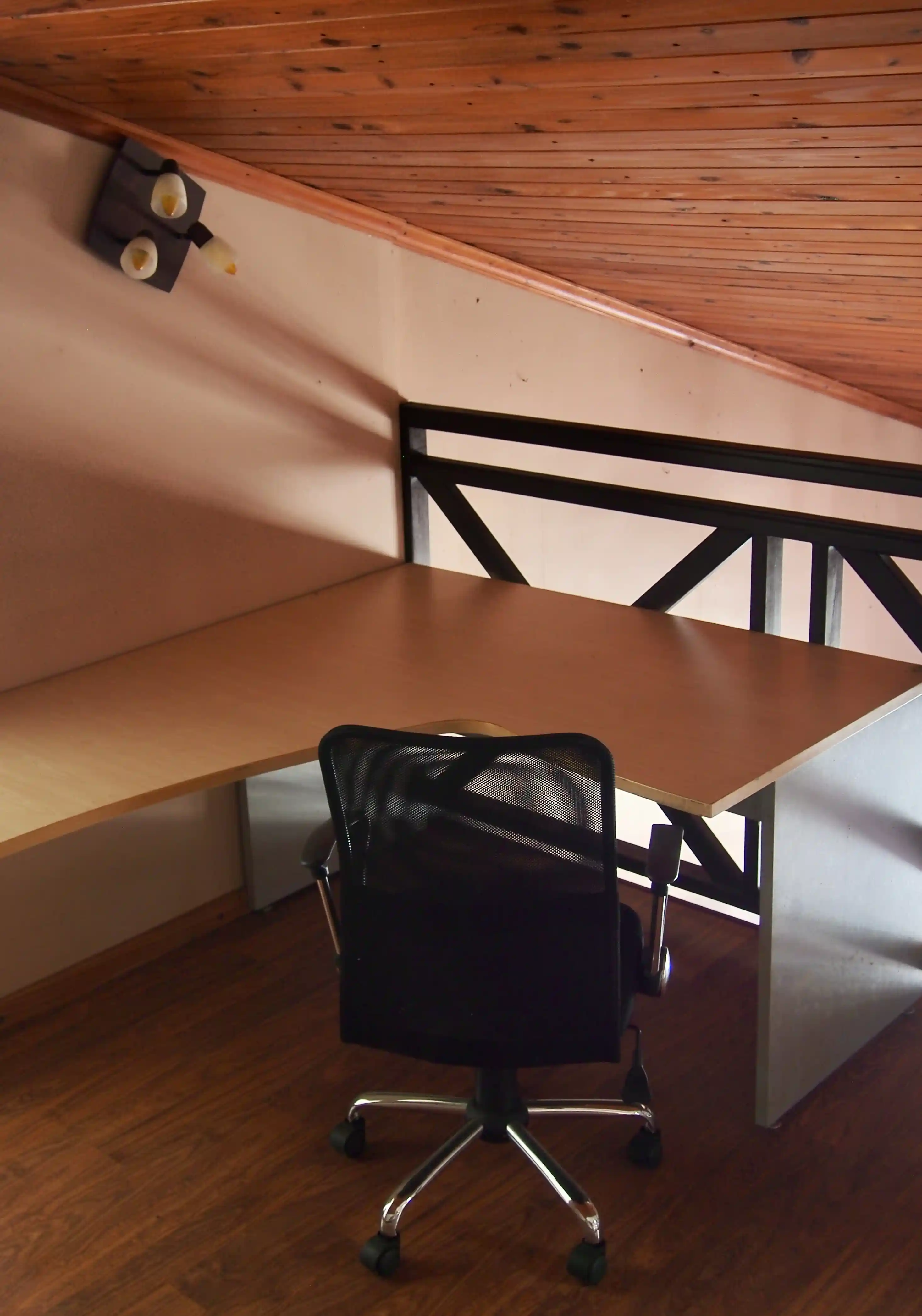 Empty black mesh office chair in front of a wooden L-shaped desk against a wall with a ceiling light fixture above.