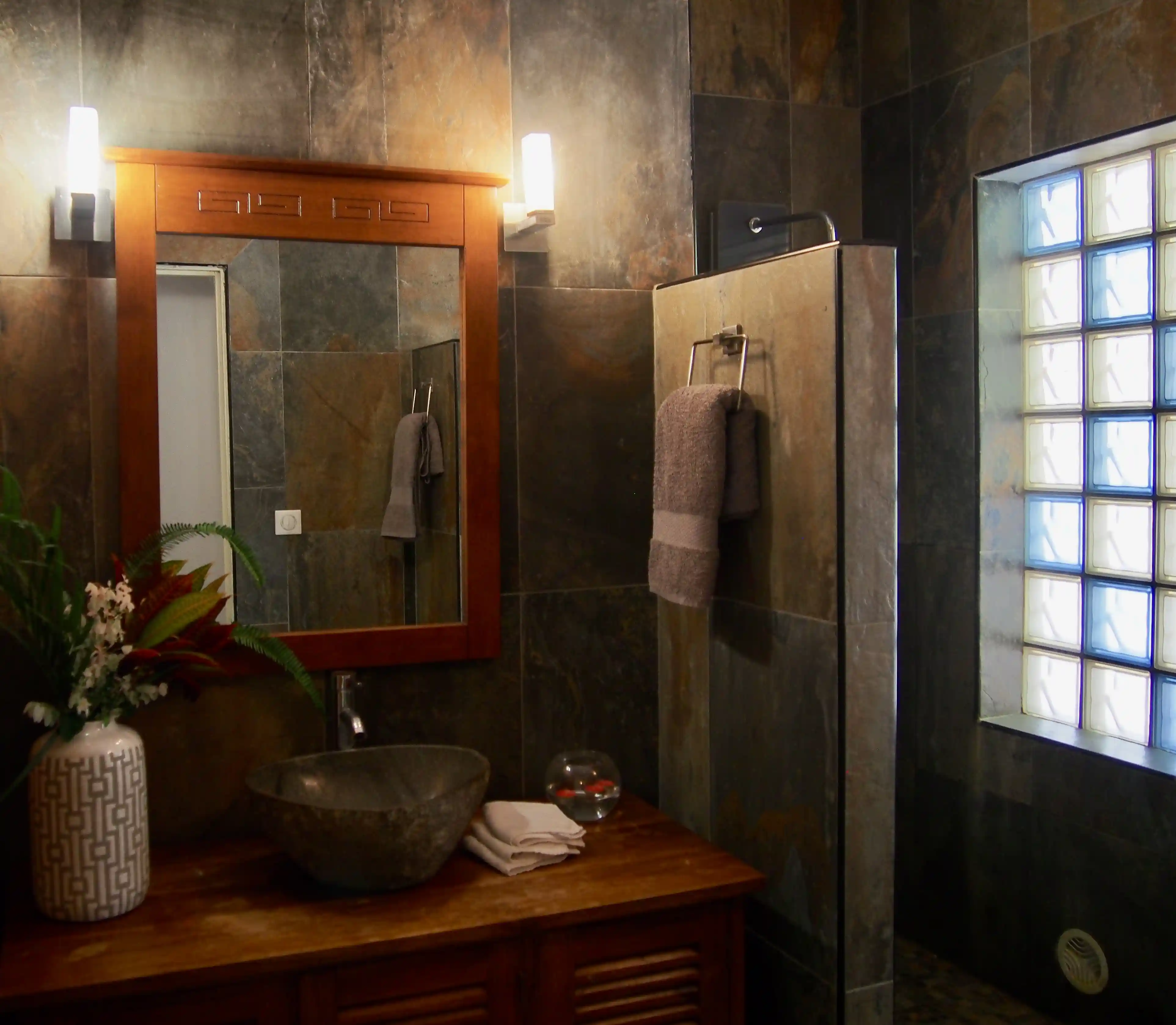 Bathroom with dark stone tiles, wooden framed mirror, stone sink basin, towel on glass partition, and frosted window blocks.