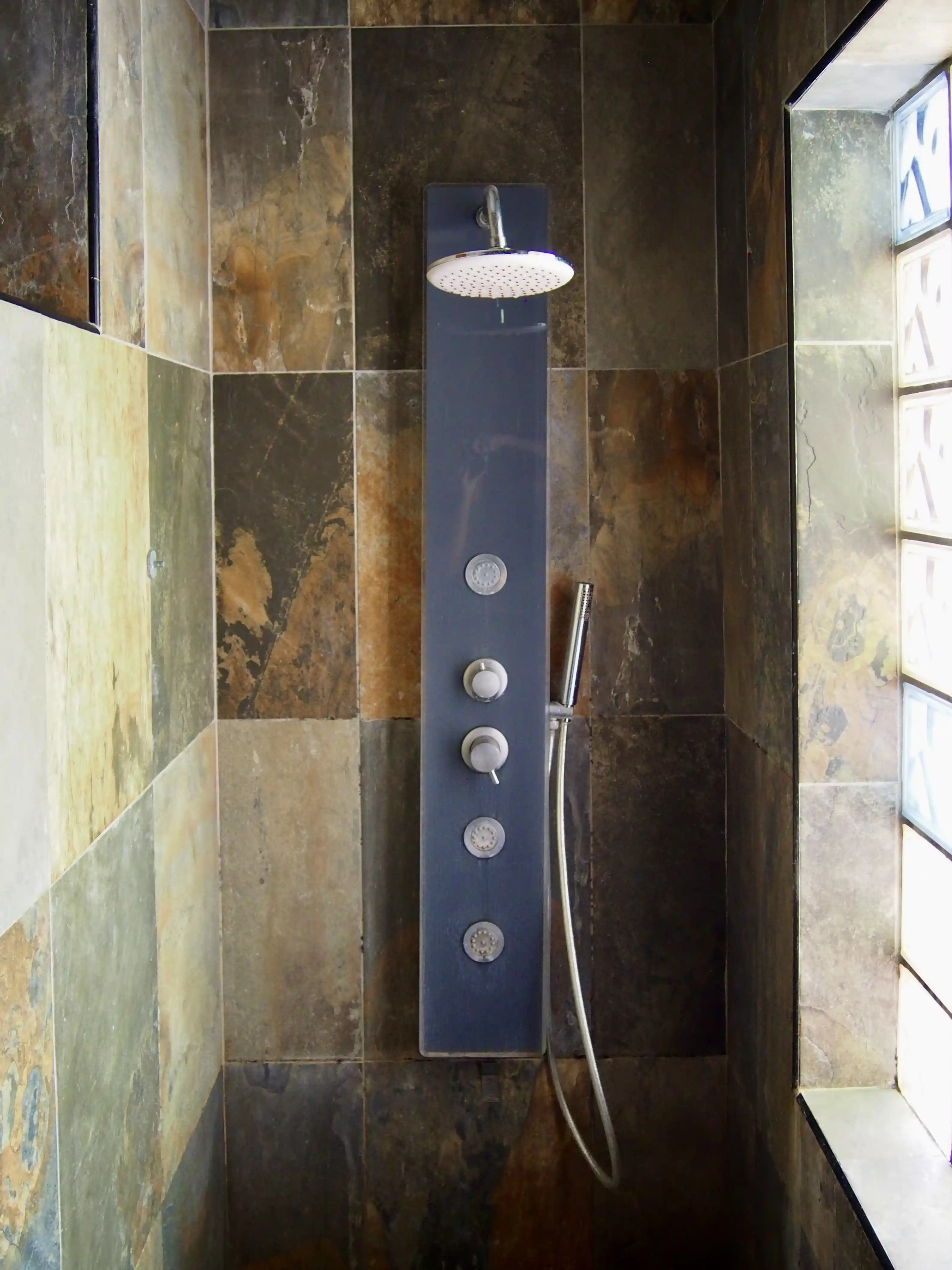 Modern shower panel with multiple controls and a handheld showerhead inside a tiled shower stall with natural stone tiles and frosted glass window.