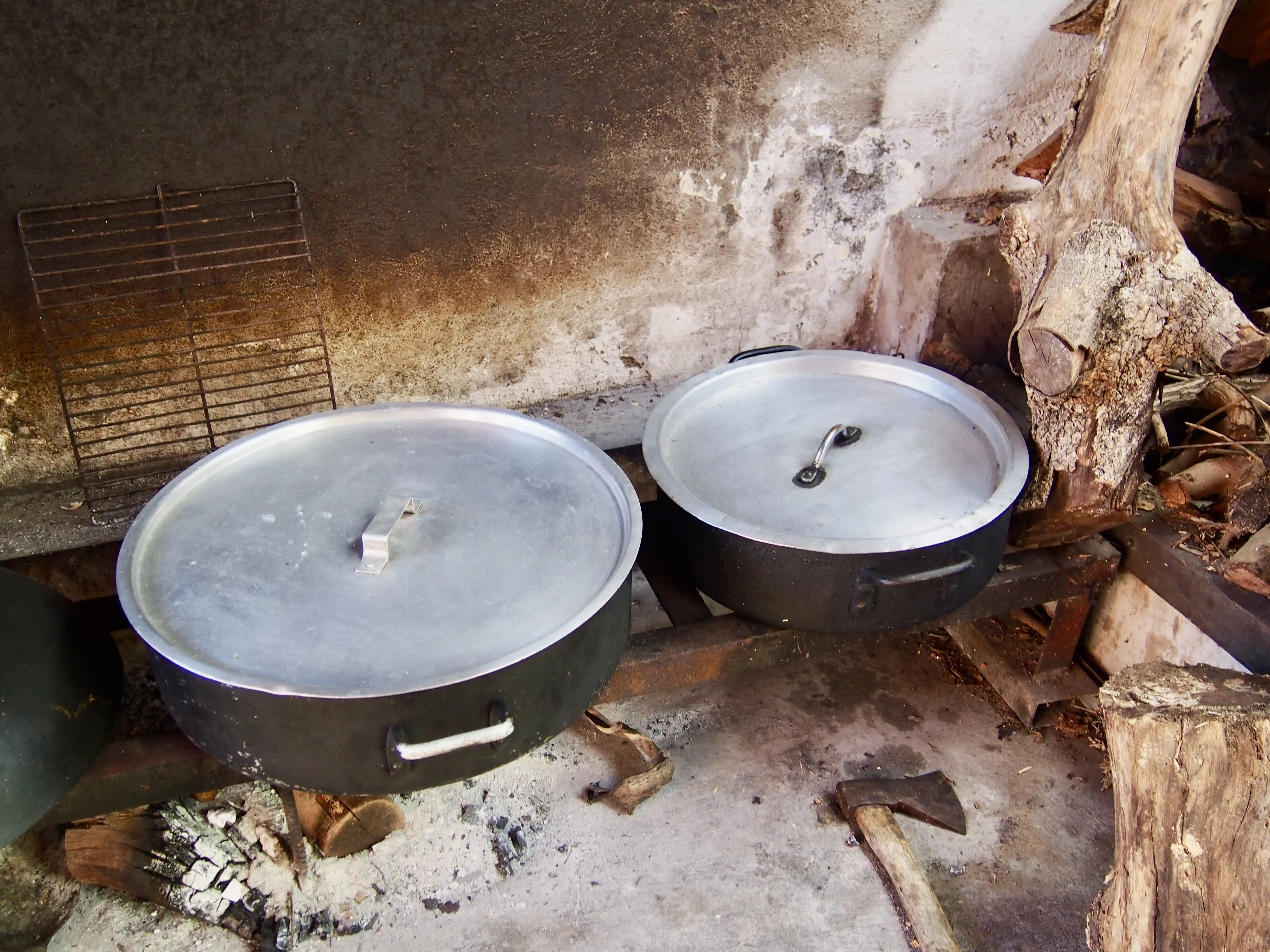Two large covered cooking pots resting on a metal frame above an outdoor fire pit with an axe and firewood nearby.